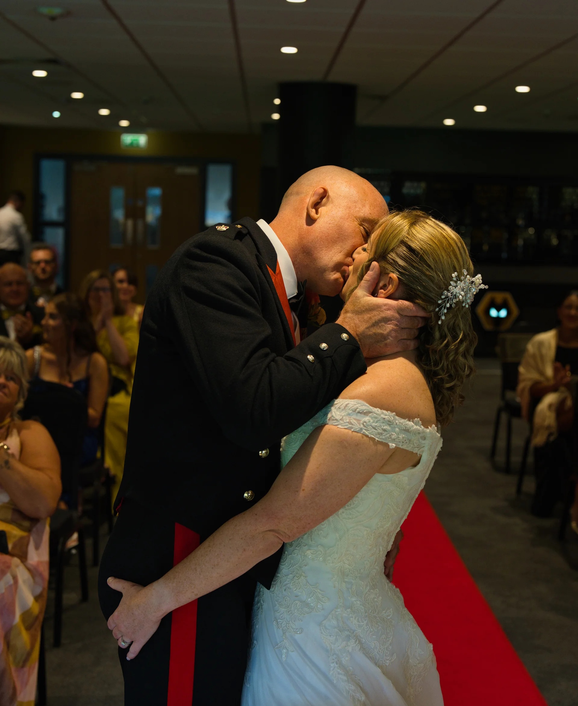 A newlywed couple shares their first kiss down a red aisle runner during a civil ceremony while guests watch in the background.