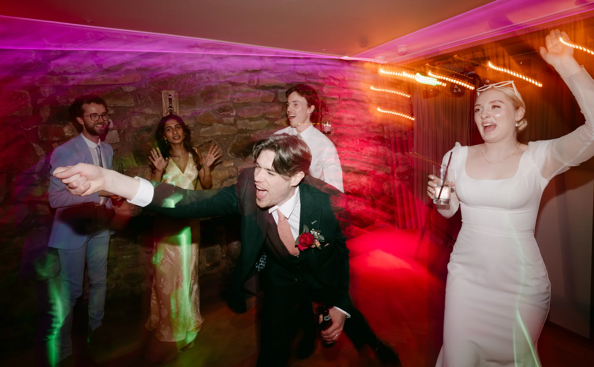 The newlyweds and wedding guests dance energetically on a packed dance floor illuminated by vibrant disco lights and direct flash photography.