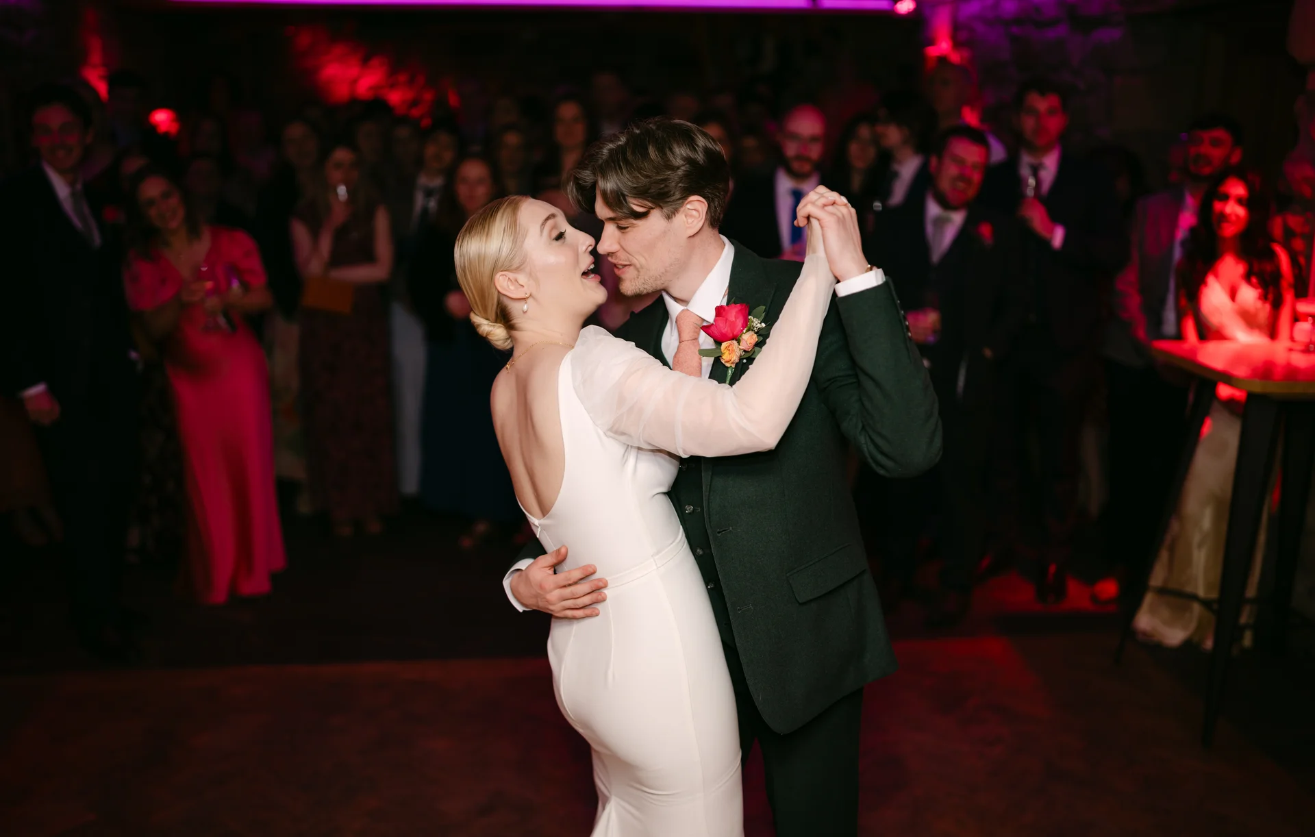 A newlywed couple shares a joyful first dance on a crowded evening dance floor, surrounded by watching guests under atmospheric red lighting.
