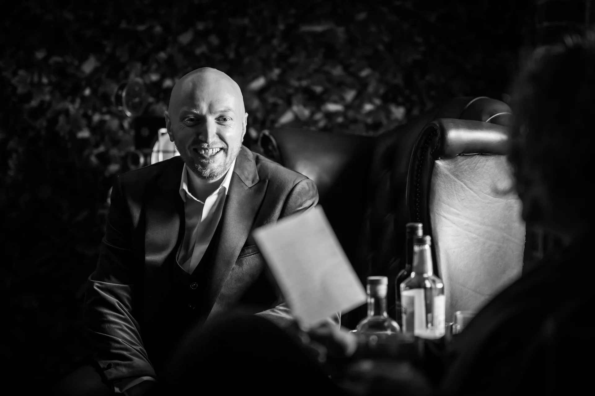 A smiling groom in a three piece suit sits in a leather armchair while listening to a speech during the wedding reception.