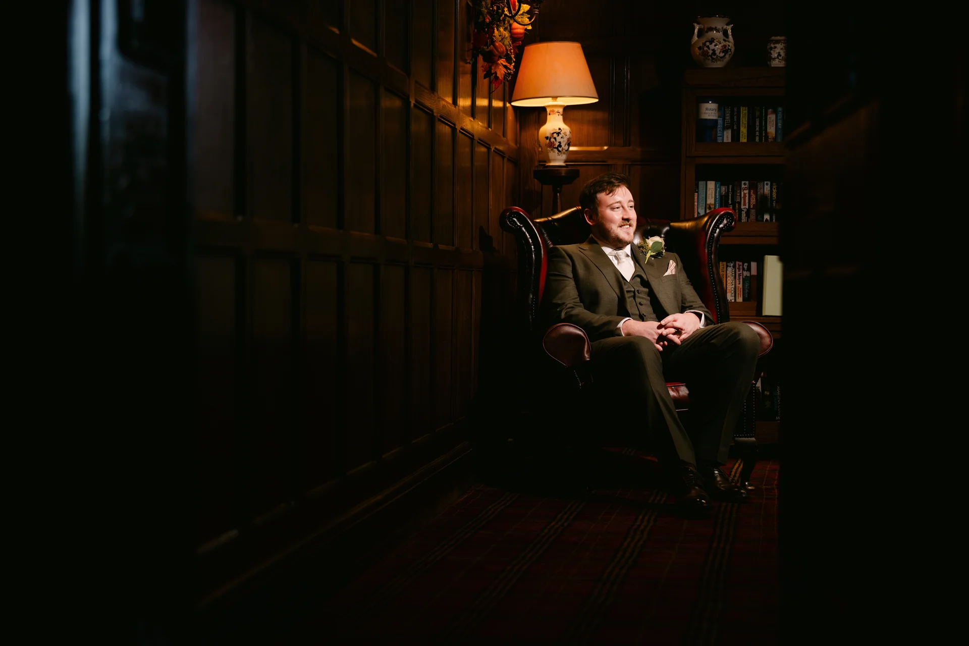 A groom in a smart green three piece suit smiles while relaxing in a leather armchair inside a wood panelled library.