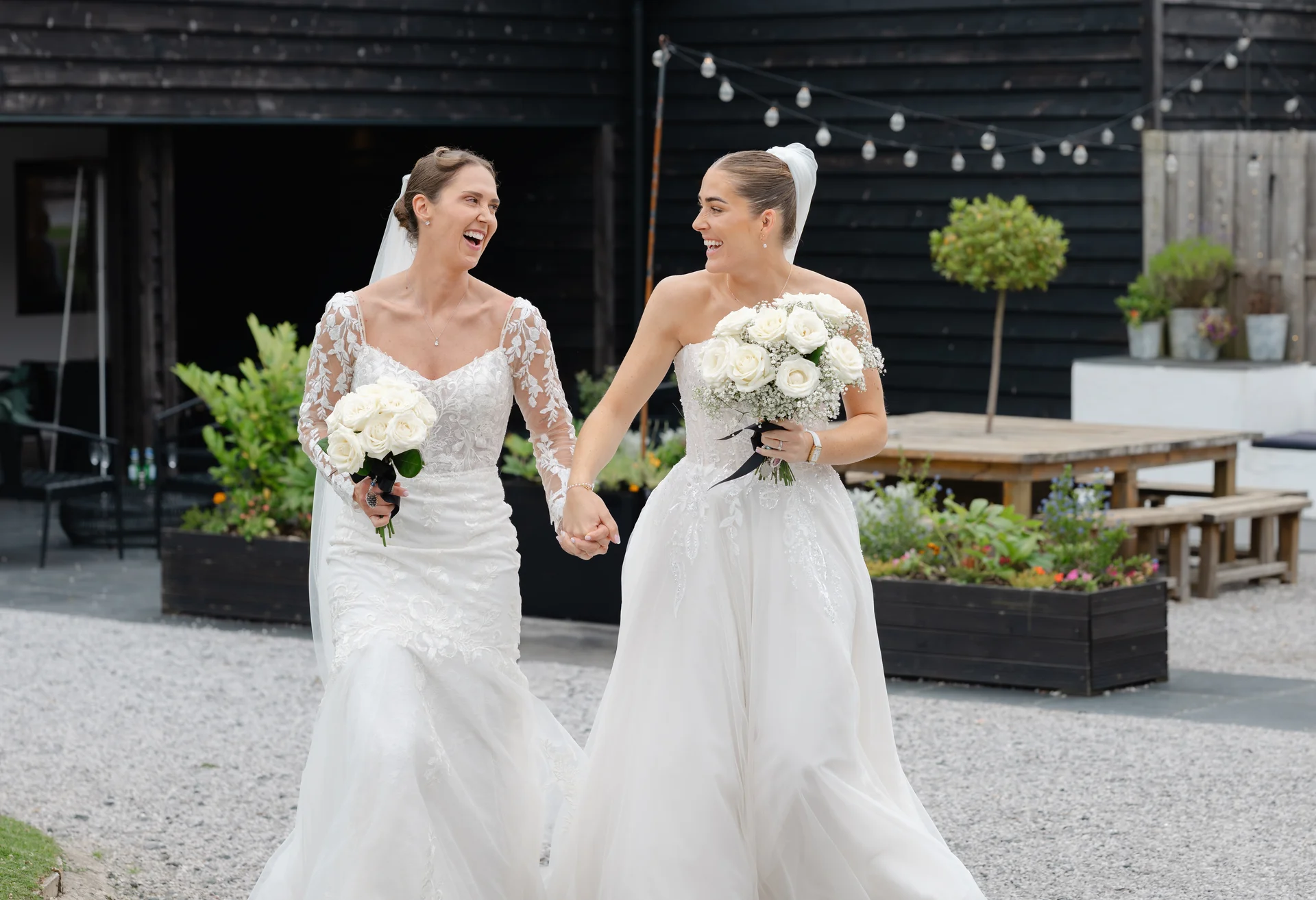 Two brides in white wedding dresses walk hand in hand outdoors in front of a black barn, laughing joyfully while holding white rose bouquets.