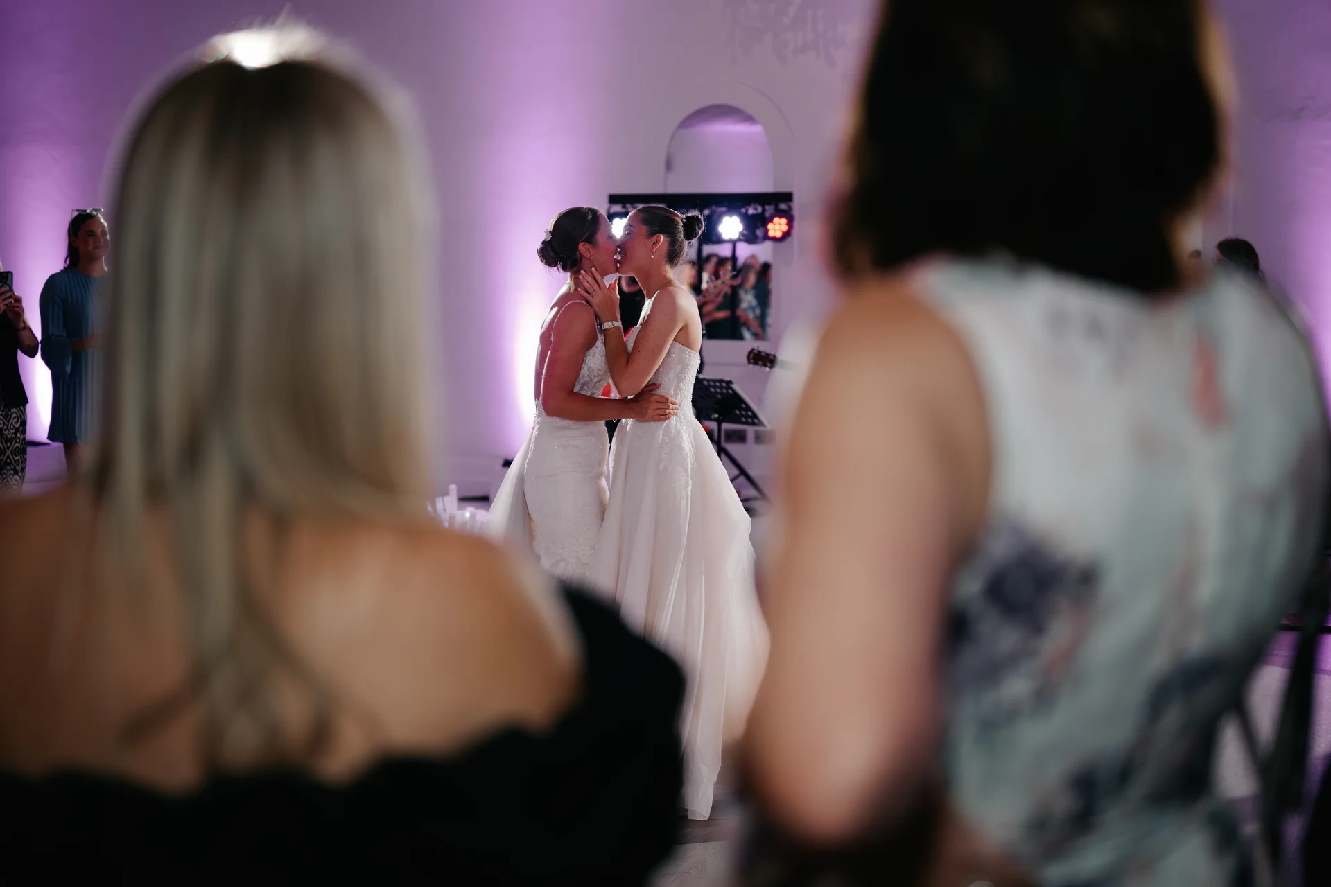 Two brides share a tender kiss on the dance floor framed by wedding guests in the foreground during an evening reception with purple lighting.
