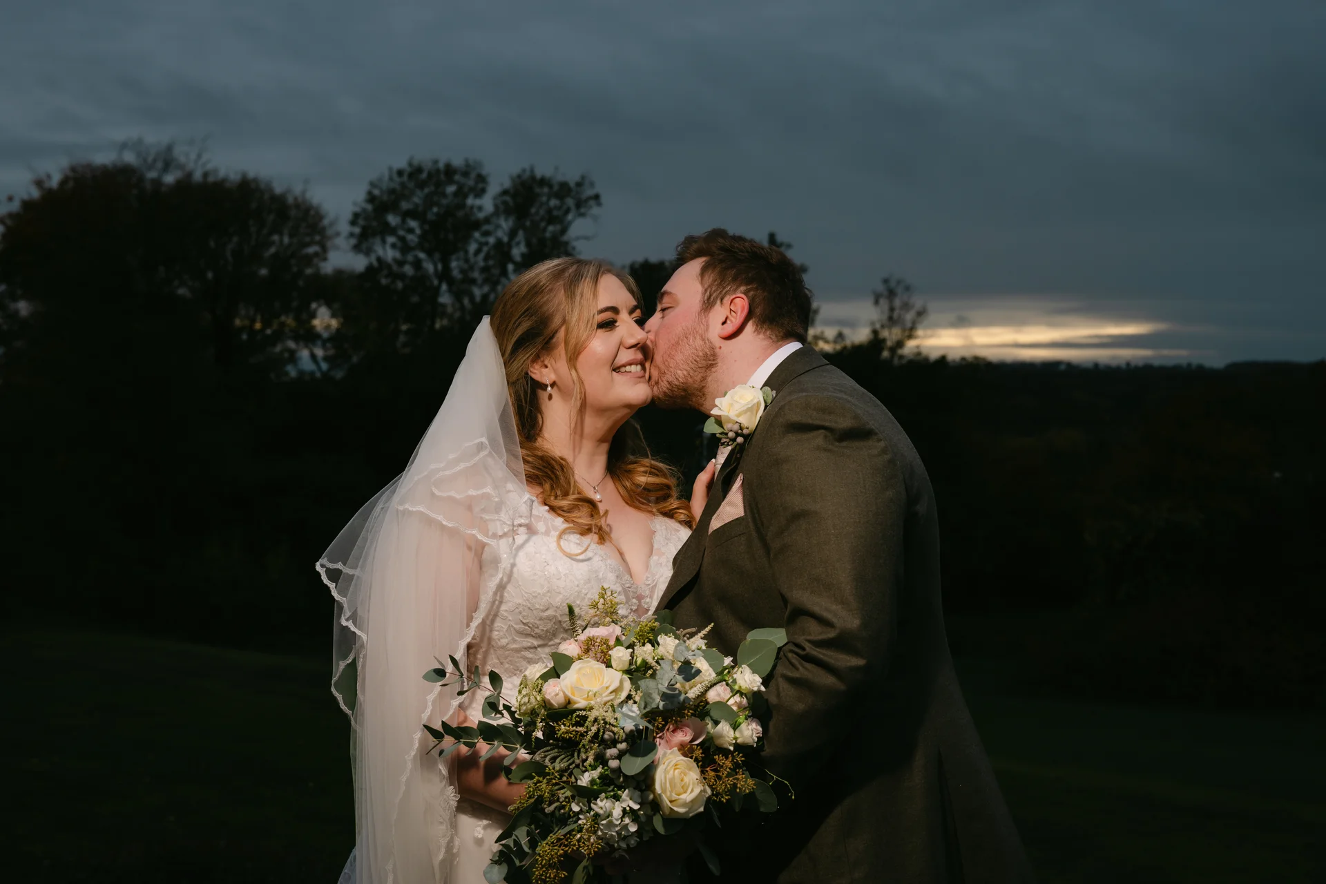 A groom gently kisses the smiling bride on the cheek outdoors during twilight, with the bride holding a lush bouquet of roses and greenery against a dramatic dark sky.