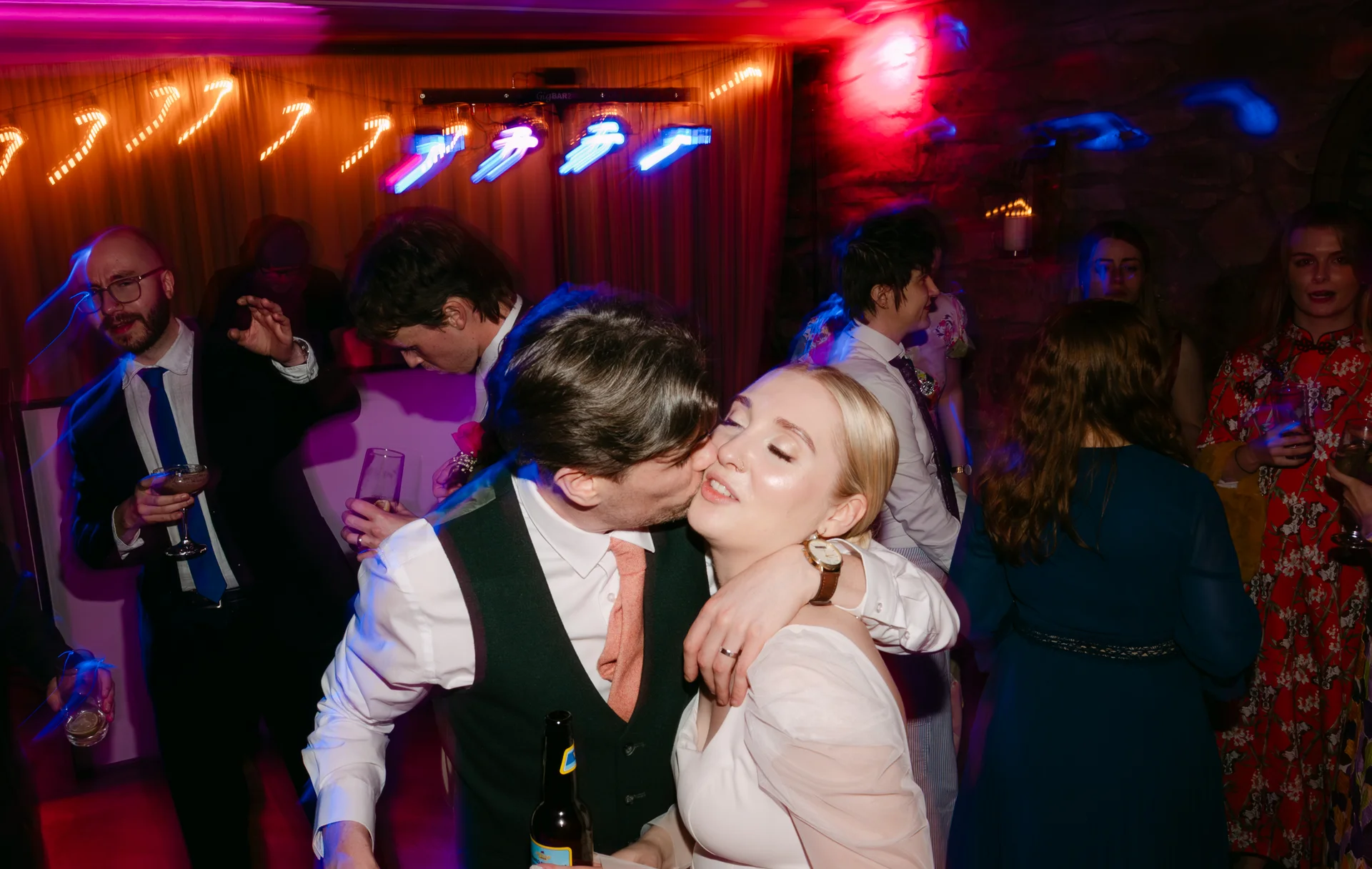 A groom kisses his bride on the cheek on a lively, brightly lit dance floor surrounded by wedding guests.