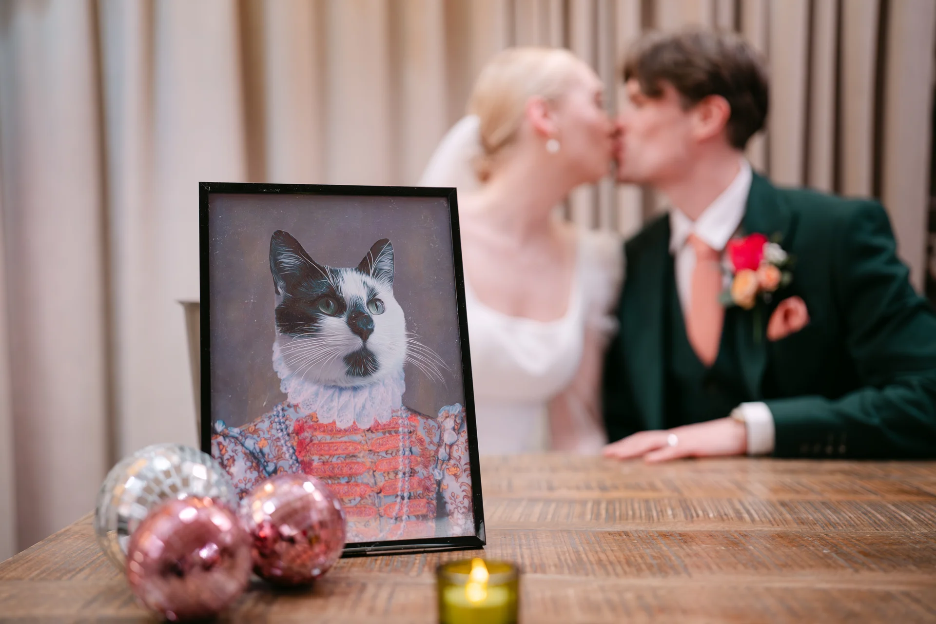 A framed portrait of a cat dressed in historic royal attire sits on a wooden table in the foreground while a bride and groom kiss softly in the softly blurred background.