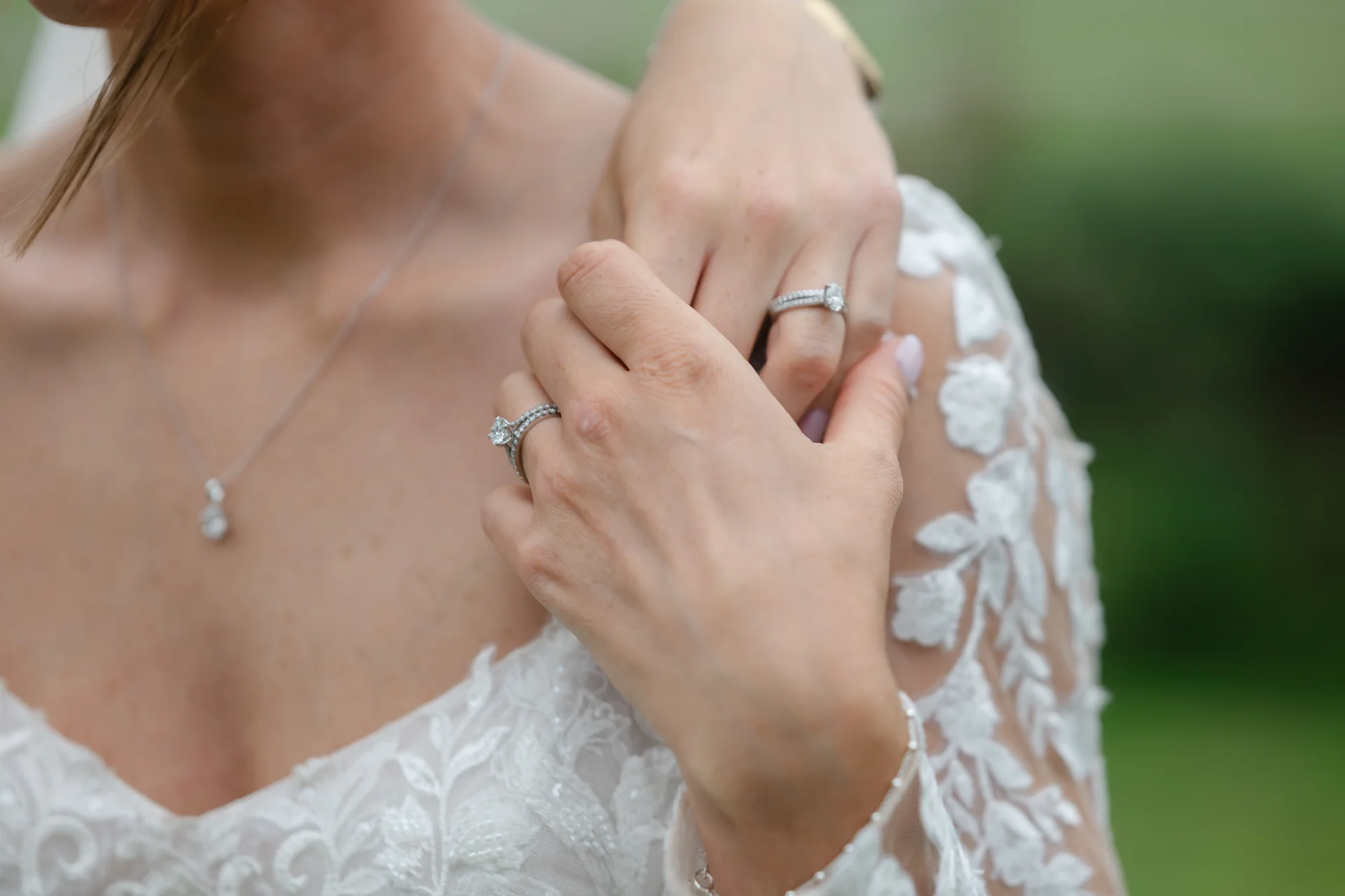 A close-up shot showing two brides holding hands, highlighting sparkling diamond engagement rings, wedding bands, and detailed lace gown sleeves.