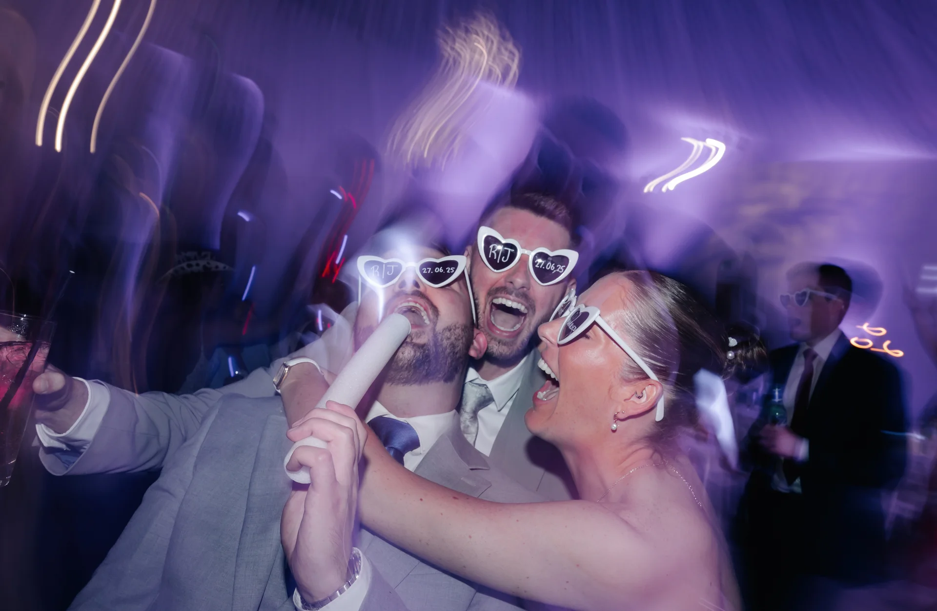 Guests wearing novelty heart-shaped sunglasses dance and sing into foam sticks on a vibrant, motion-blurred dance floor illuminated by purple party lights.