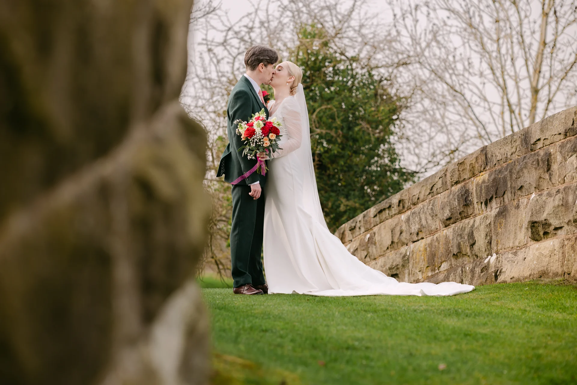 A newlywed couple shares a tender kiss next to a stone wall, with the bride holding a vibrant bouquet containing red roses.