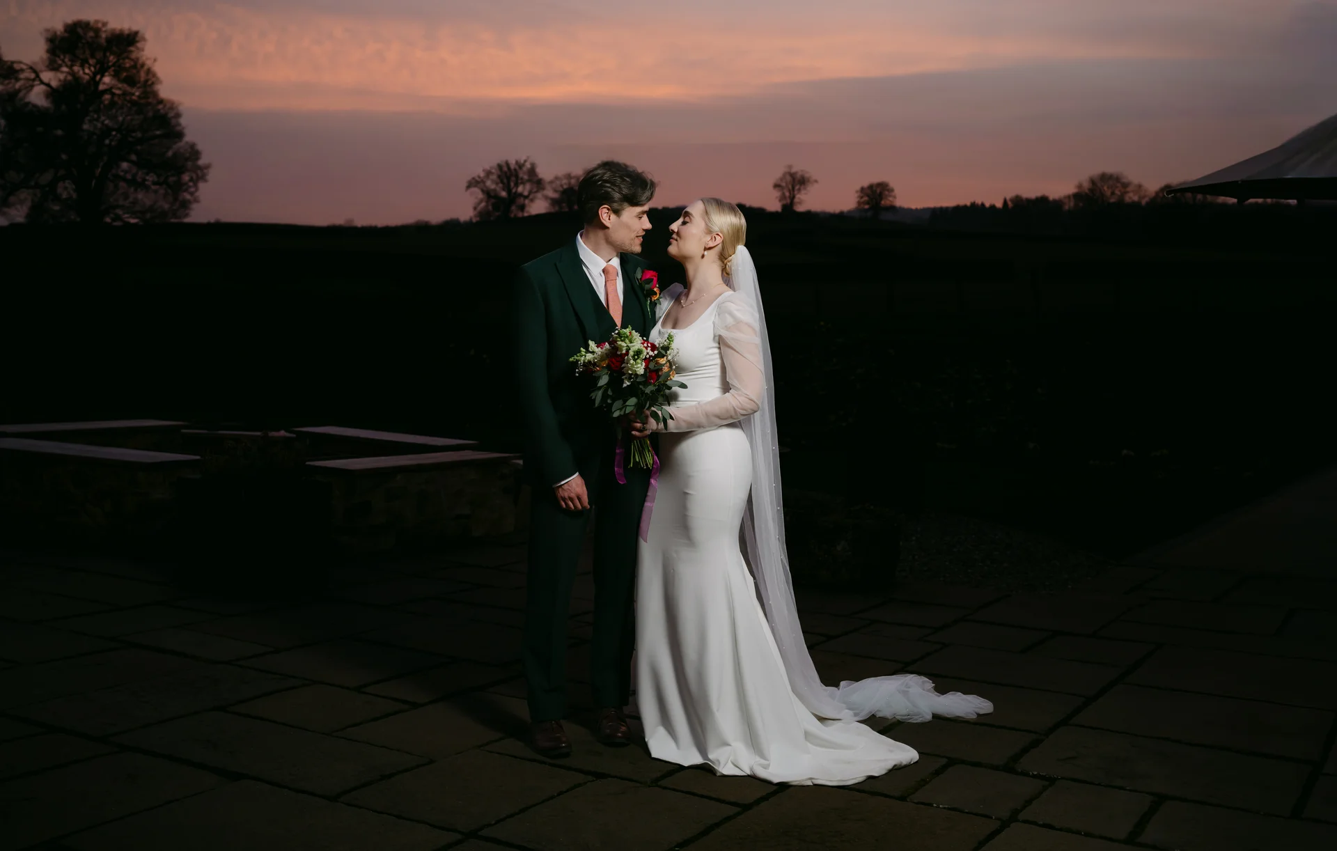 A newlywed couple shares a tender gaze outdoors on a stone patio against a dramatic sunset sky with rolling hills in the distance.