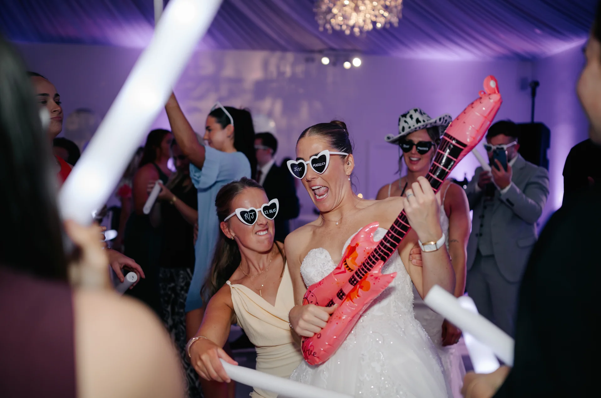 The bride energetically plays an inflatable guitar on a packed dance floor while wearing heart-shaped sunglasses, surrounded by cheering guests holding glow sticks at an evening wedding reception.