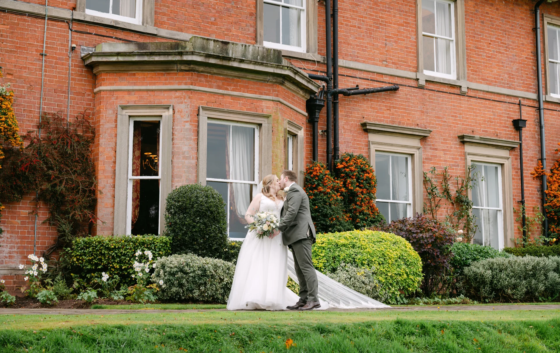 A newlywed couple shares a tender kiss in front of a grand red brick manor house surrounded by lush green gardens.