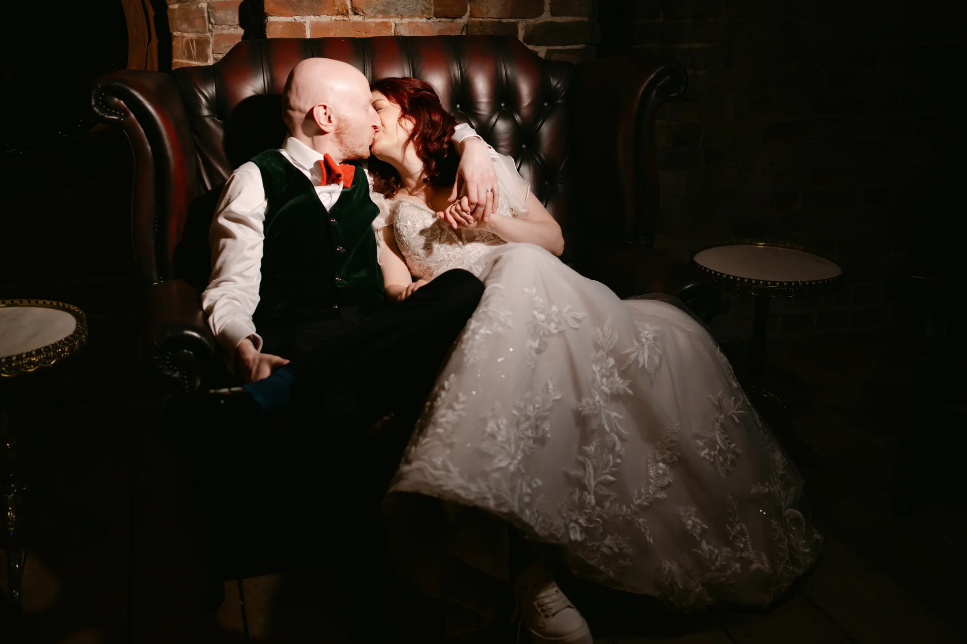 A bride with red hair and a groom wearing a green velvet vest share a tender kiss while lounging together on a dark leather Chesterfield sofa in a moody room with exposed brick walls.
