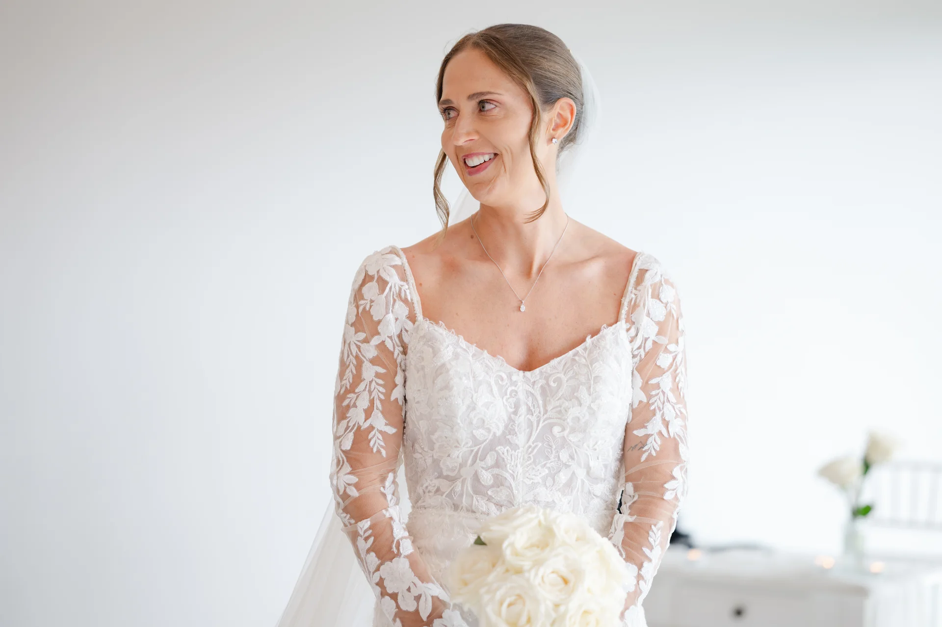 A smiling bride in a long-sleeve lace wedding gown holds a white rose bouquet during her portrait session.