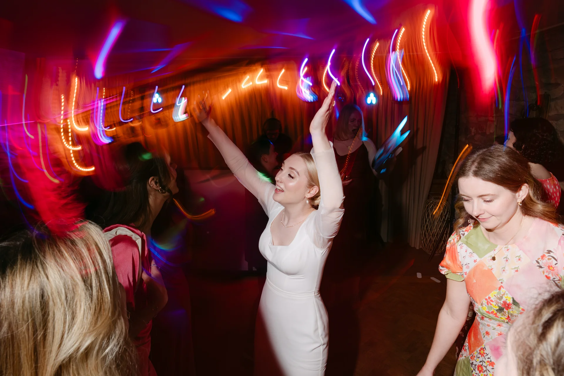 A bride in a white long-sleeved dress dances with her arms raised on a vibrant, motion-blurred dance floor illuminated by colourful disco lights.