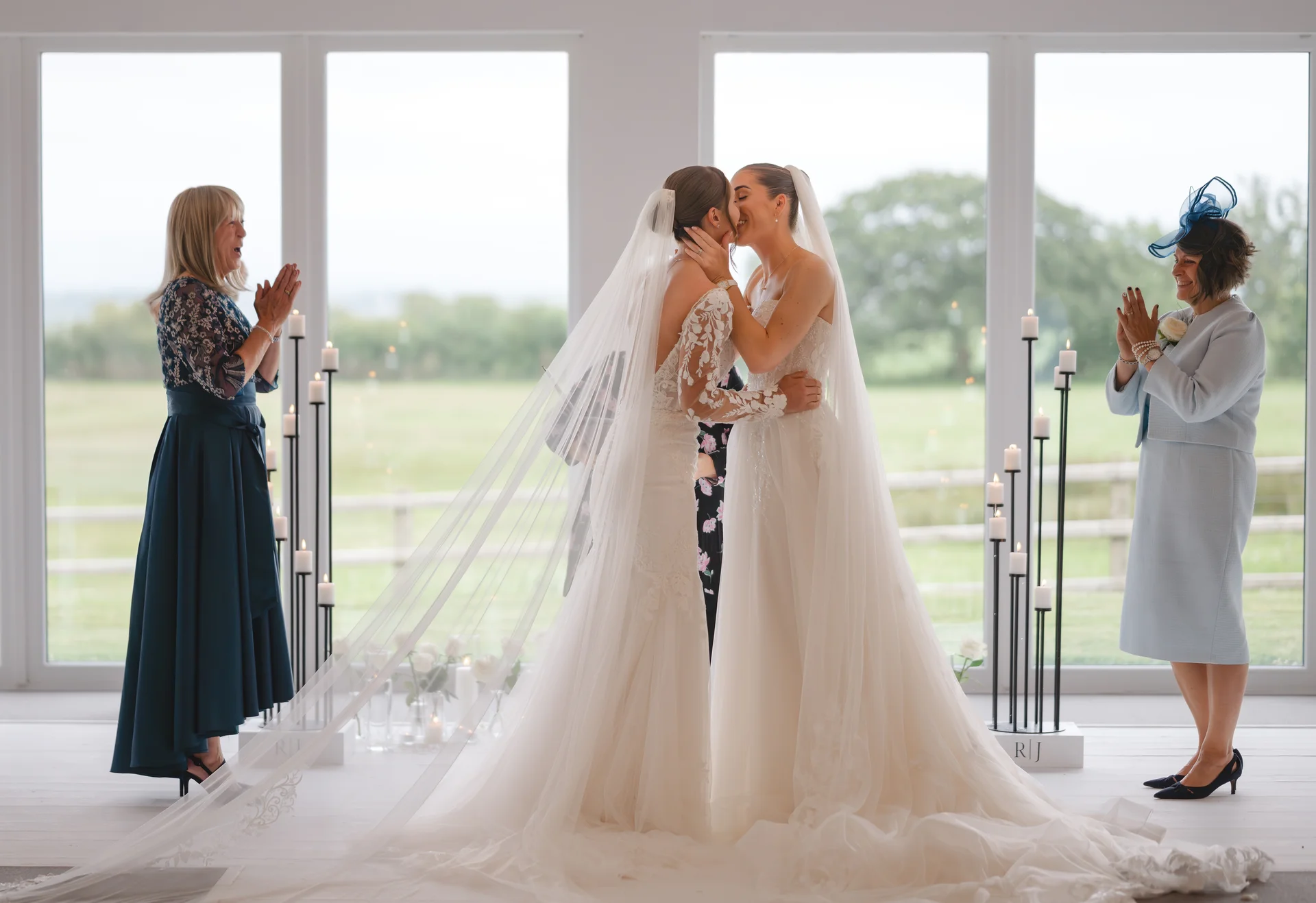 Two brides share their first kiss during a modern indoor civil ceremony while family members look on and applaud against a scenic window backdrop.