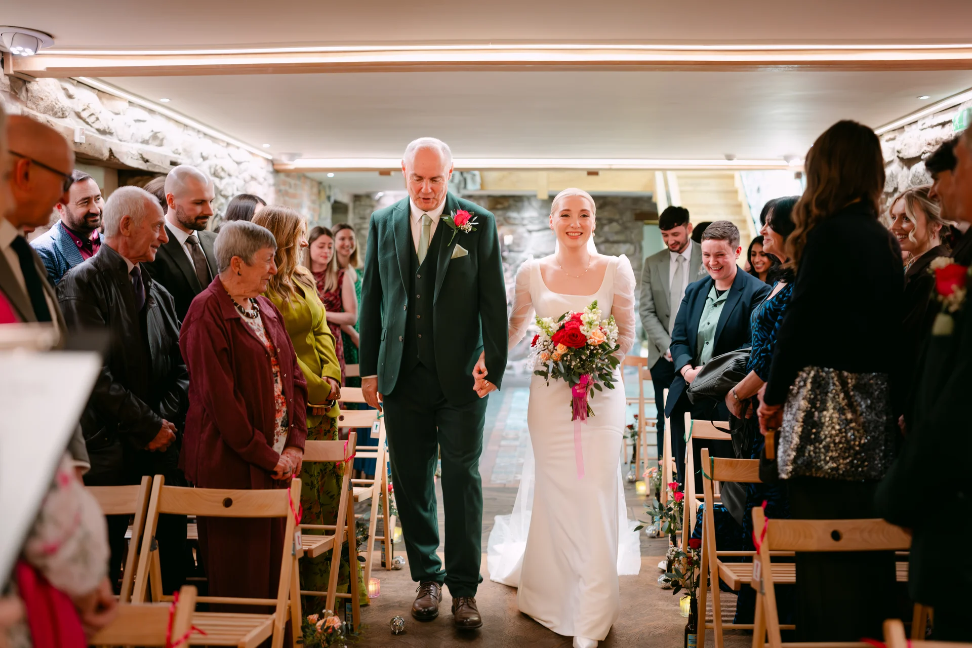 A smiling bride holding a colourful bouquet walks down the aisle arm in arm with her father, flanked by seated wedding guests inside a rustic stone barn venue.