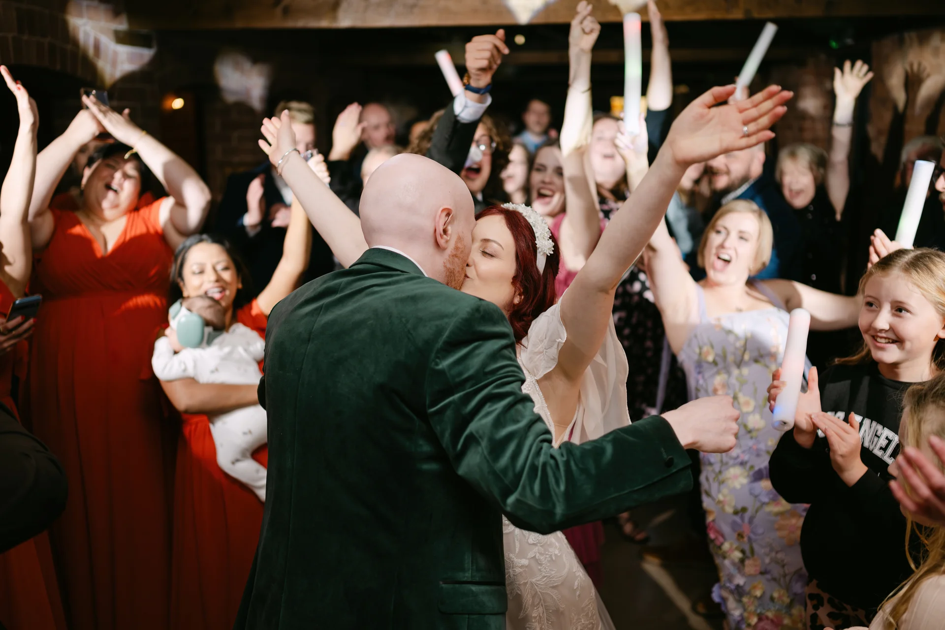 A groom in a green velvet jacket and a bride kissing on a lively dance floor as wedding guests cheer with glow sticks.