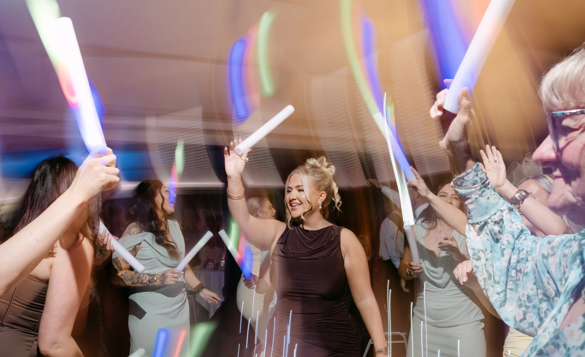 Guests on a lively dance floor wave glowing foam sticks in the air with dynamic motion blur and colorful party lighting.