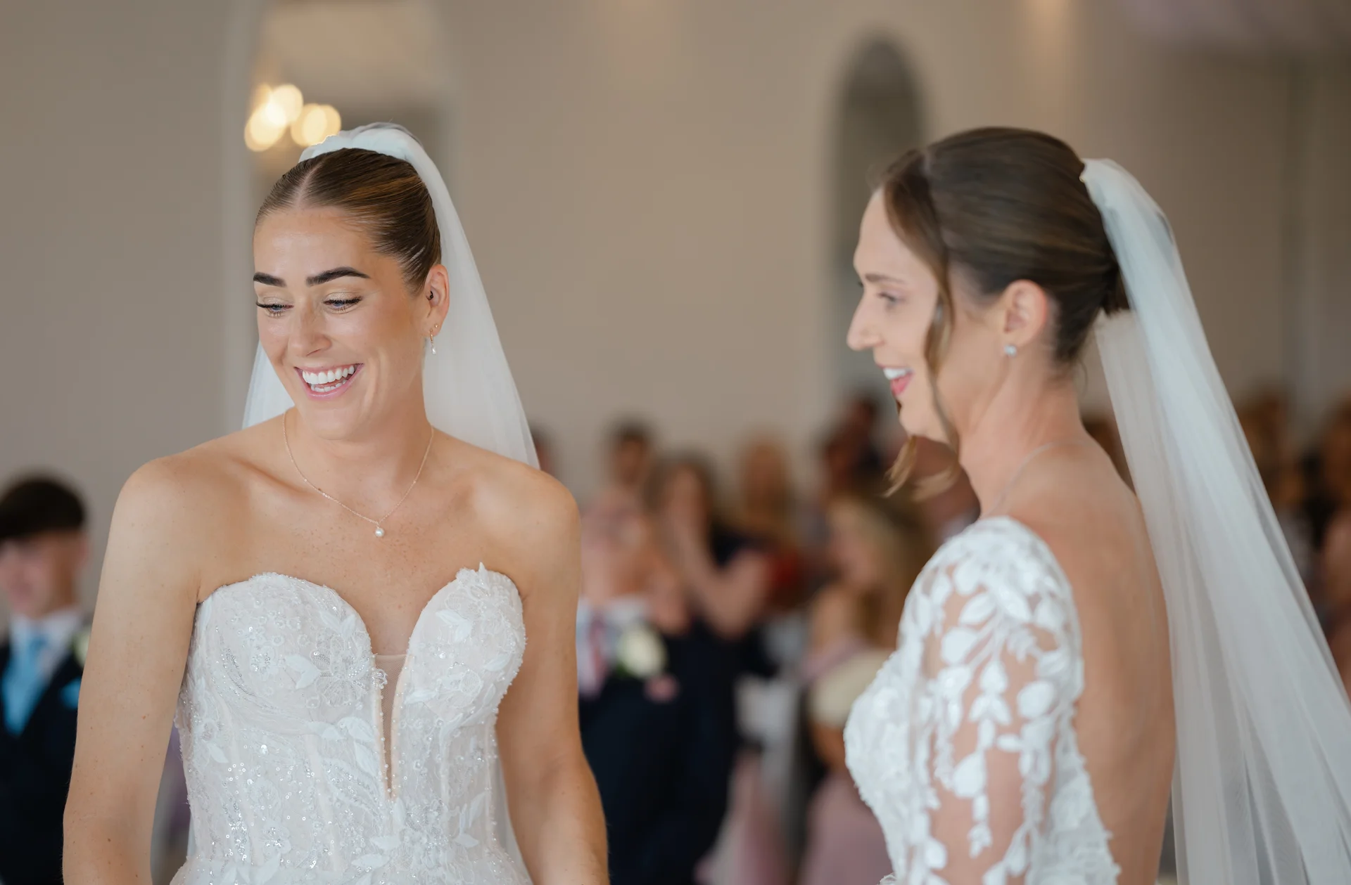 Two smiling brides in detailed white wedding gowns stand together during their indoor ceremony while guests look on softly in the background.