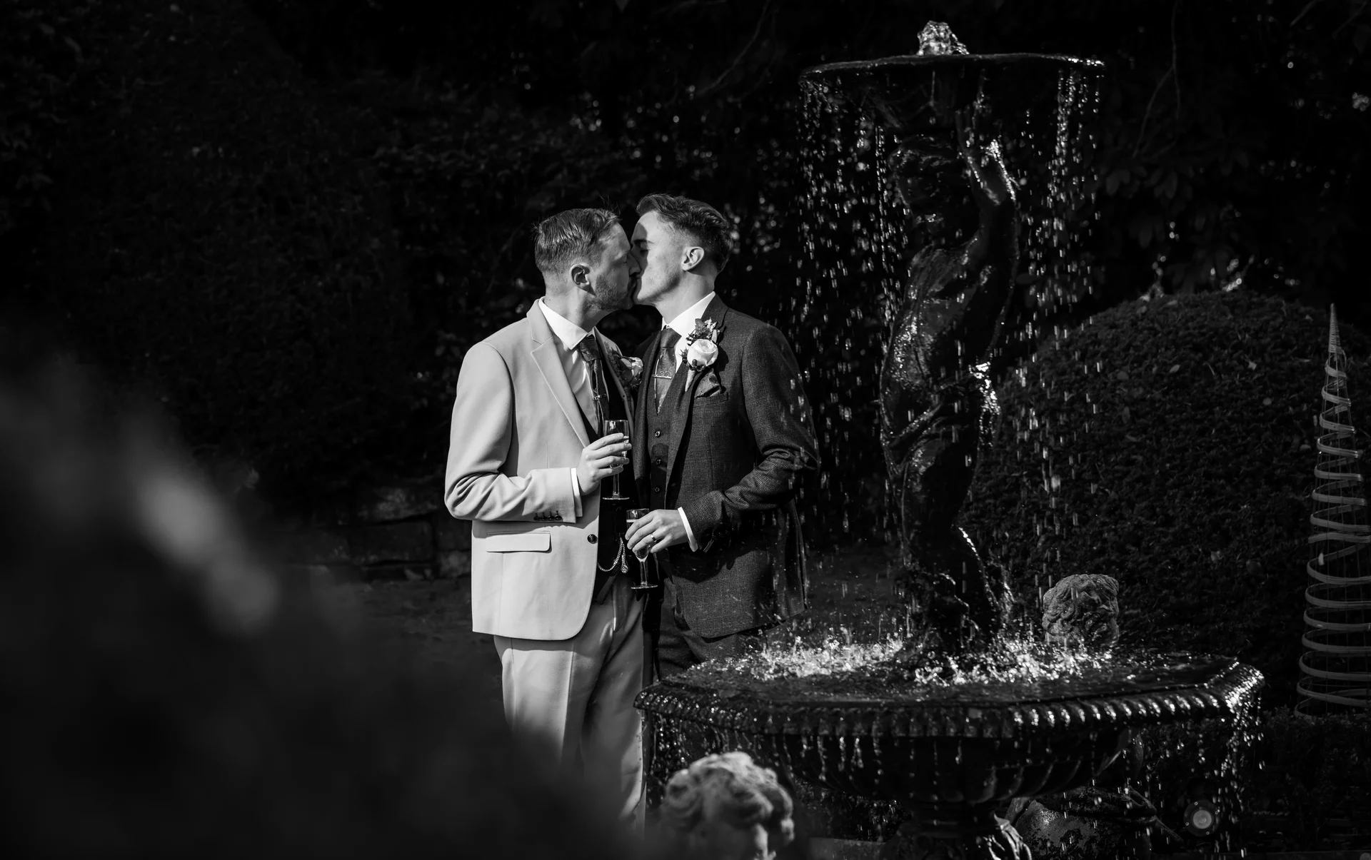 Two grooms share a tender kiss outdoors beside a cascading garden fountain in this striking black and white photograph.