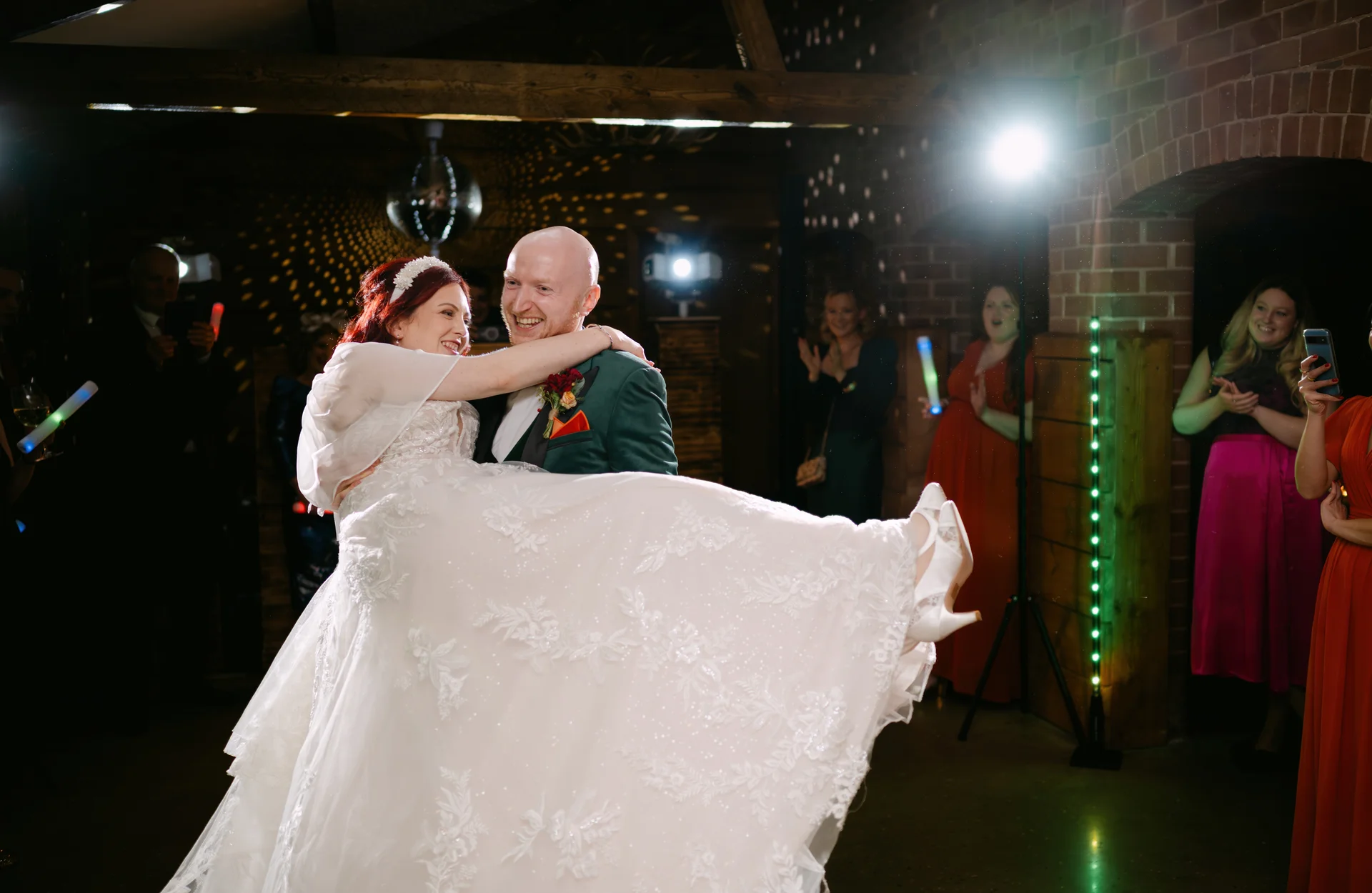 The groom joyfully lifts his bride into the air during their first dance inside a rustic brick barn venue, surrounded by cheering guests and glowing party lights.