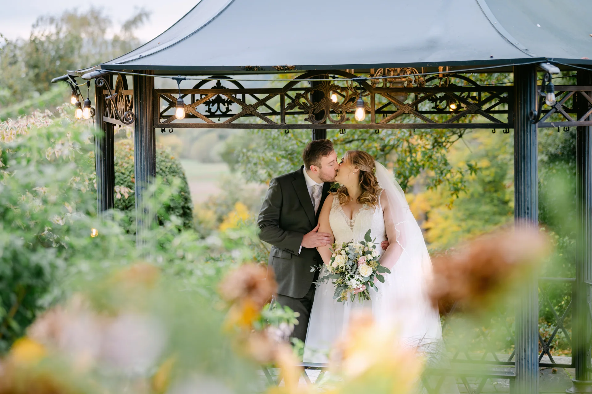A newlywed couple shares a tender kiss inside an ornate black metal gazebo surrounded by autumn foliage and soft fairy lights.