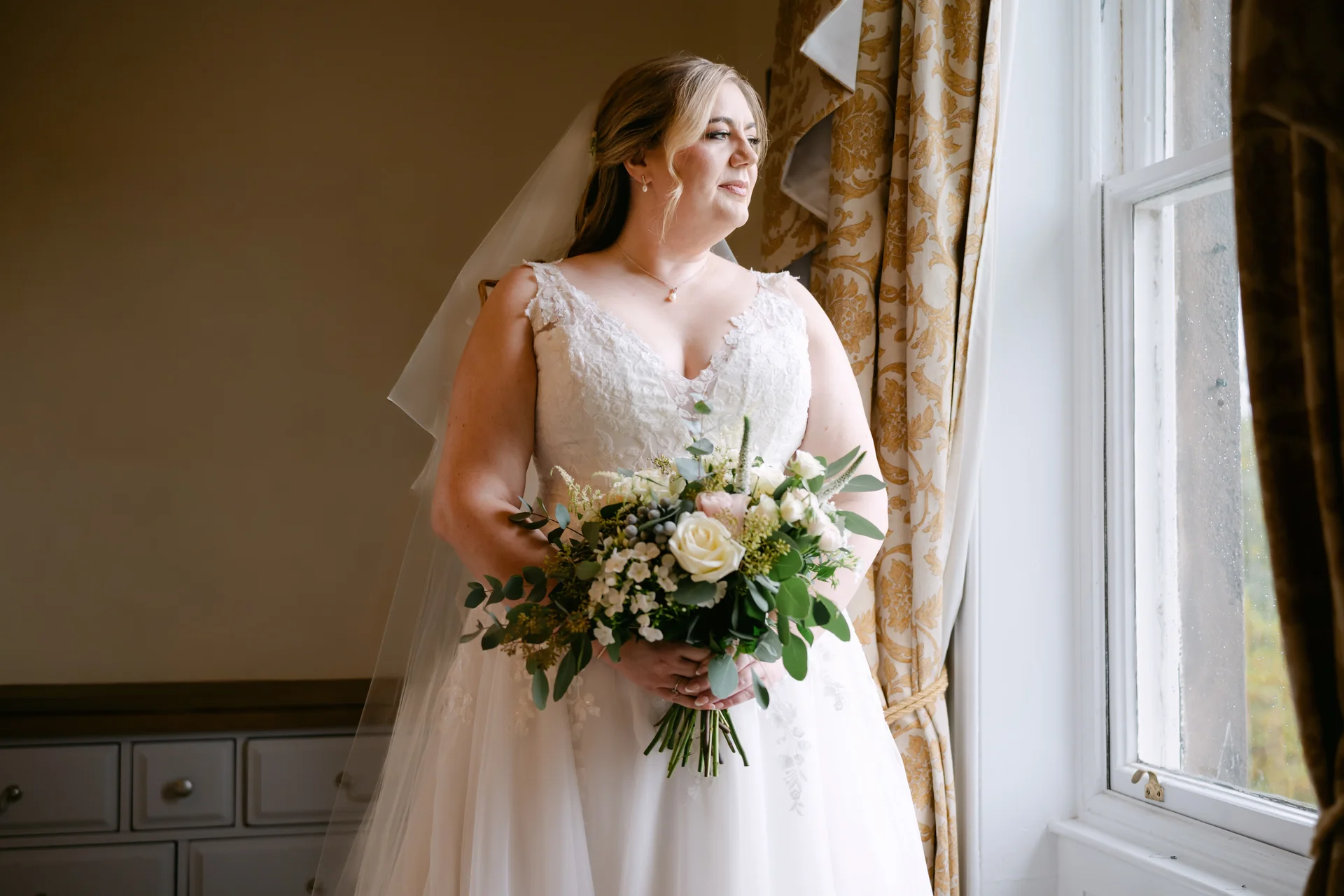 A beautiful bride stands by a window in natural light, wearing a lace a-line wedding dress and holding a hand-tied bouquet of roses and greenery.
