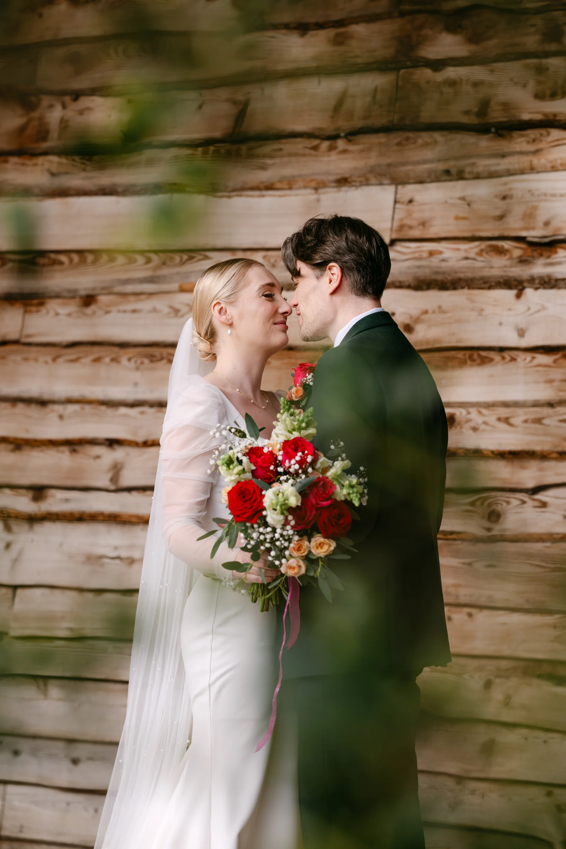 A bride and groom share a tender moment nose to nose against a rustic wood wall, with the bride holding a vibrant red rose bouquet.