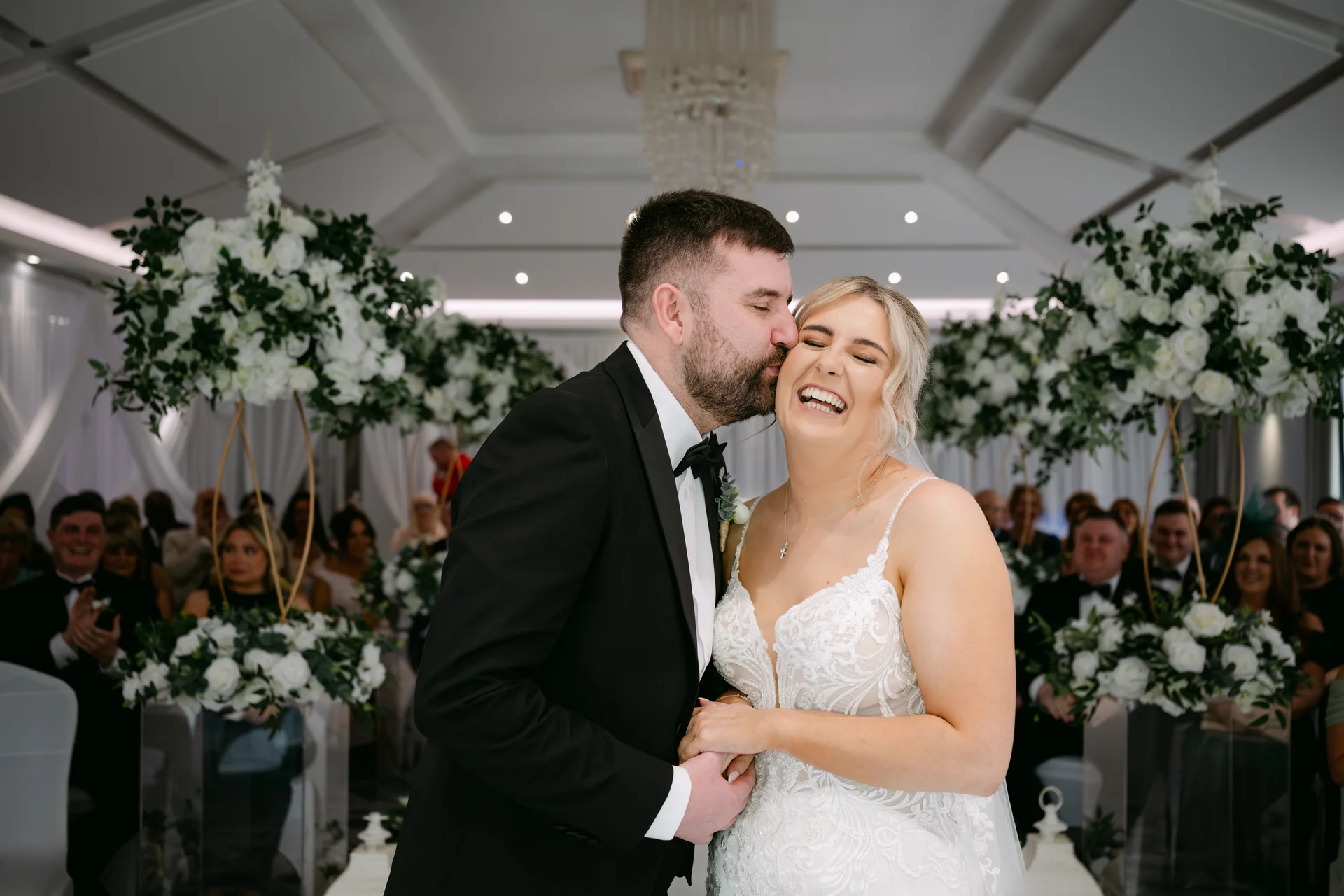 The groom kisses the smiling bride on the cheek during their indoor wedding ceremony framed by white floral arches.