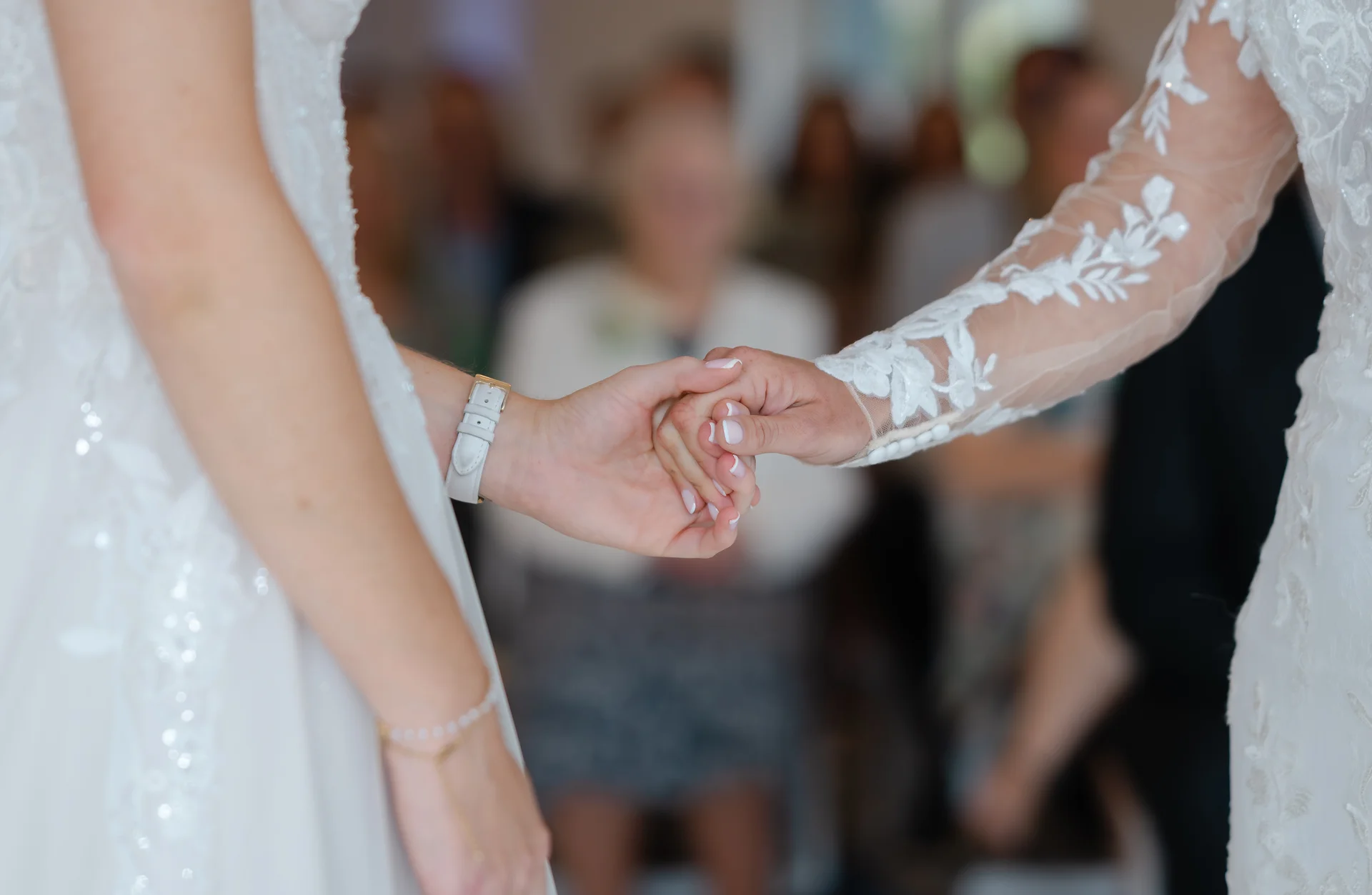 A close-up shot captures the tender moment of two brides holding hands during their wedding ceremony, showcasing delicate lace sleeves and French manicured nails.