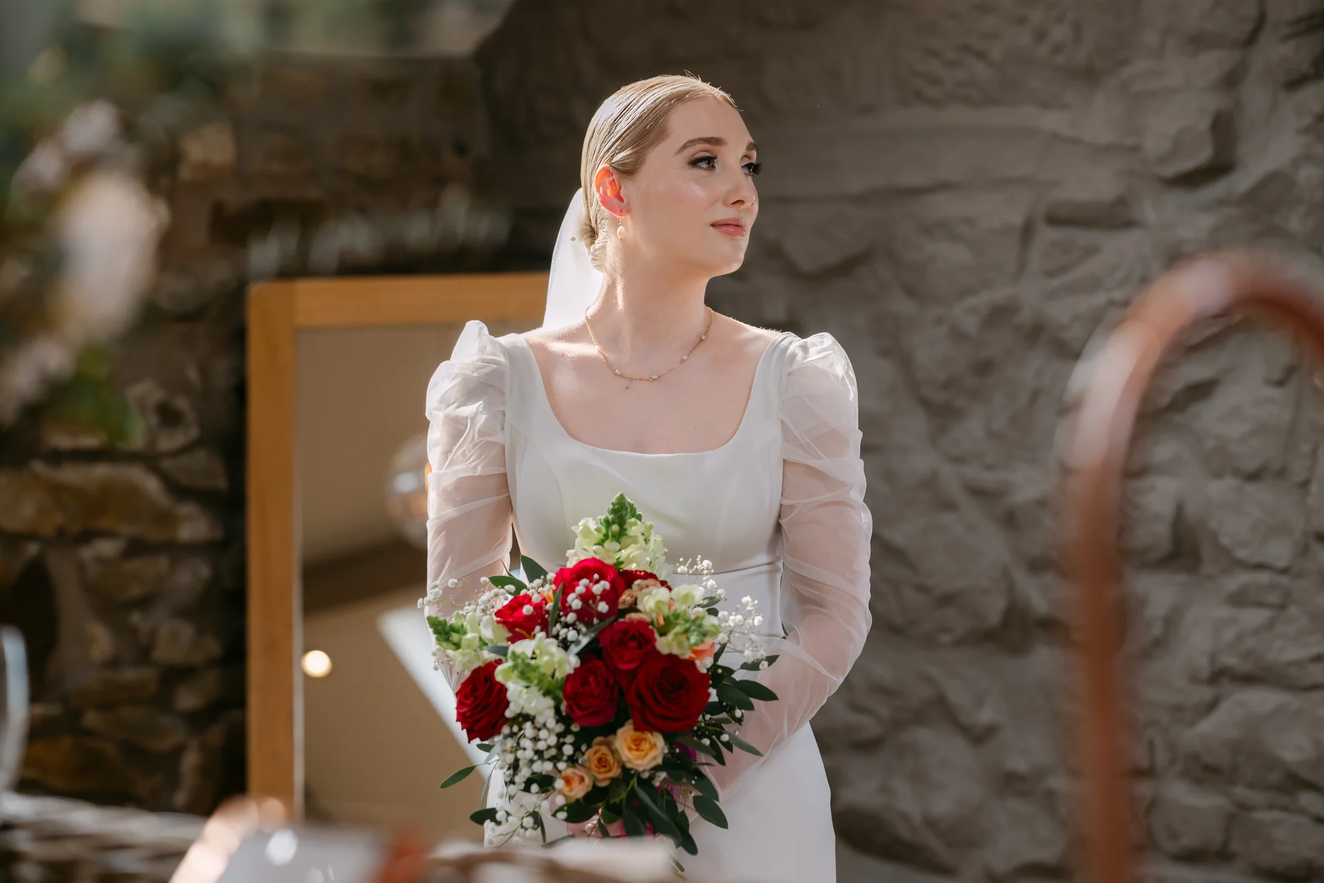 A stunning solo bride holding a vibrant red rose bouquet stands against an exposed stone wall, wearing a long-sleeved gown with sheer puff sleeves and a square neckline.