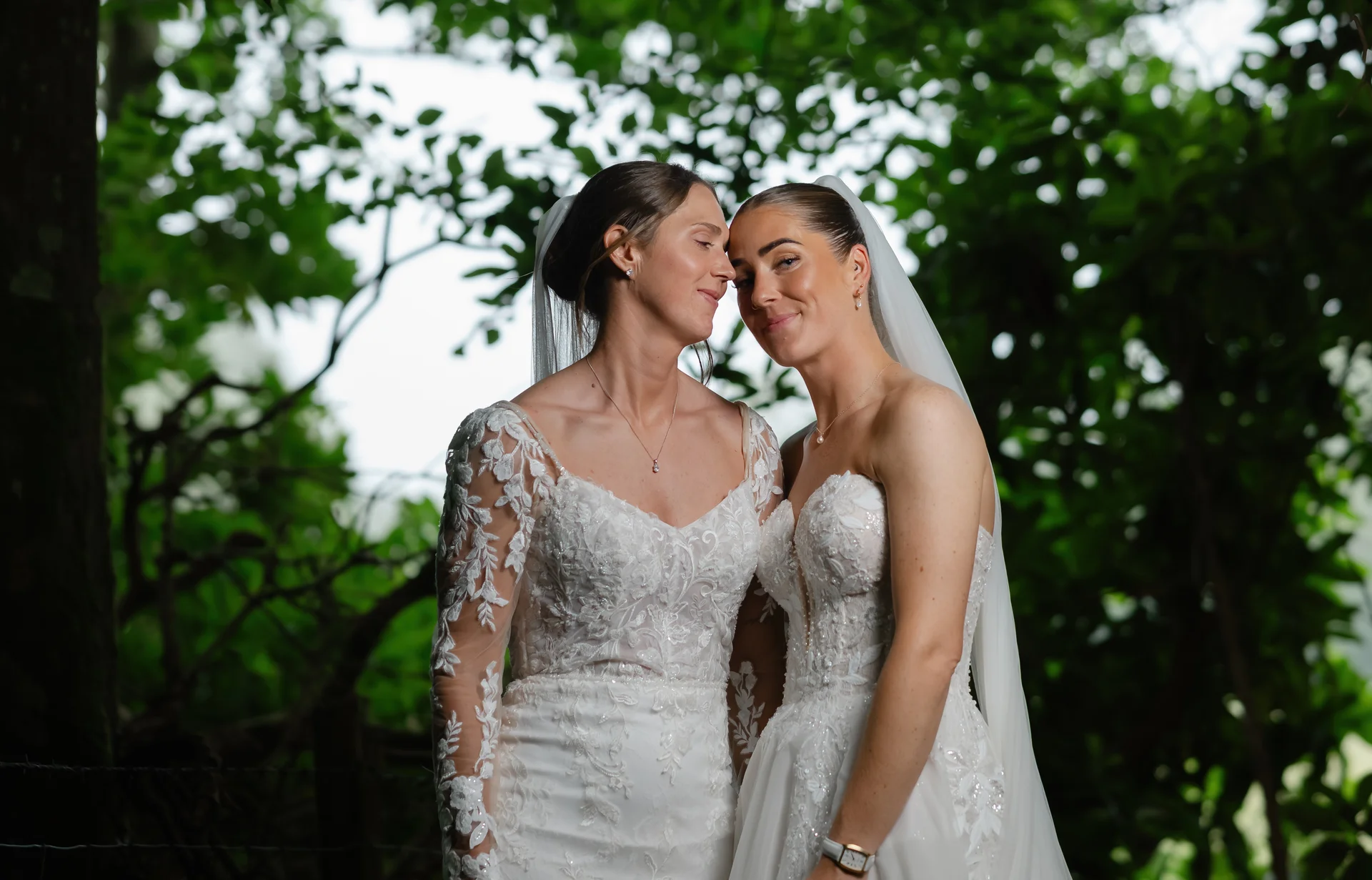 Two brides pose intimately together outdoors, framed by lush green foliage, wearing elegant white wedding gowns with intricate lace detailing and veils.