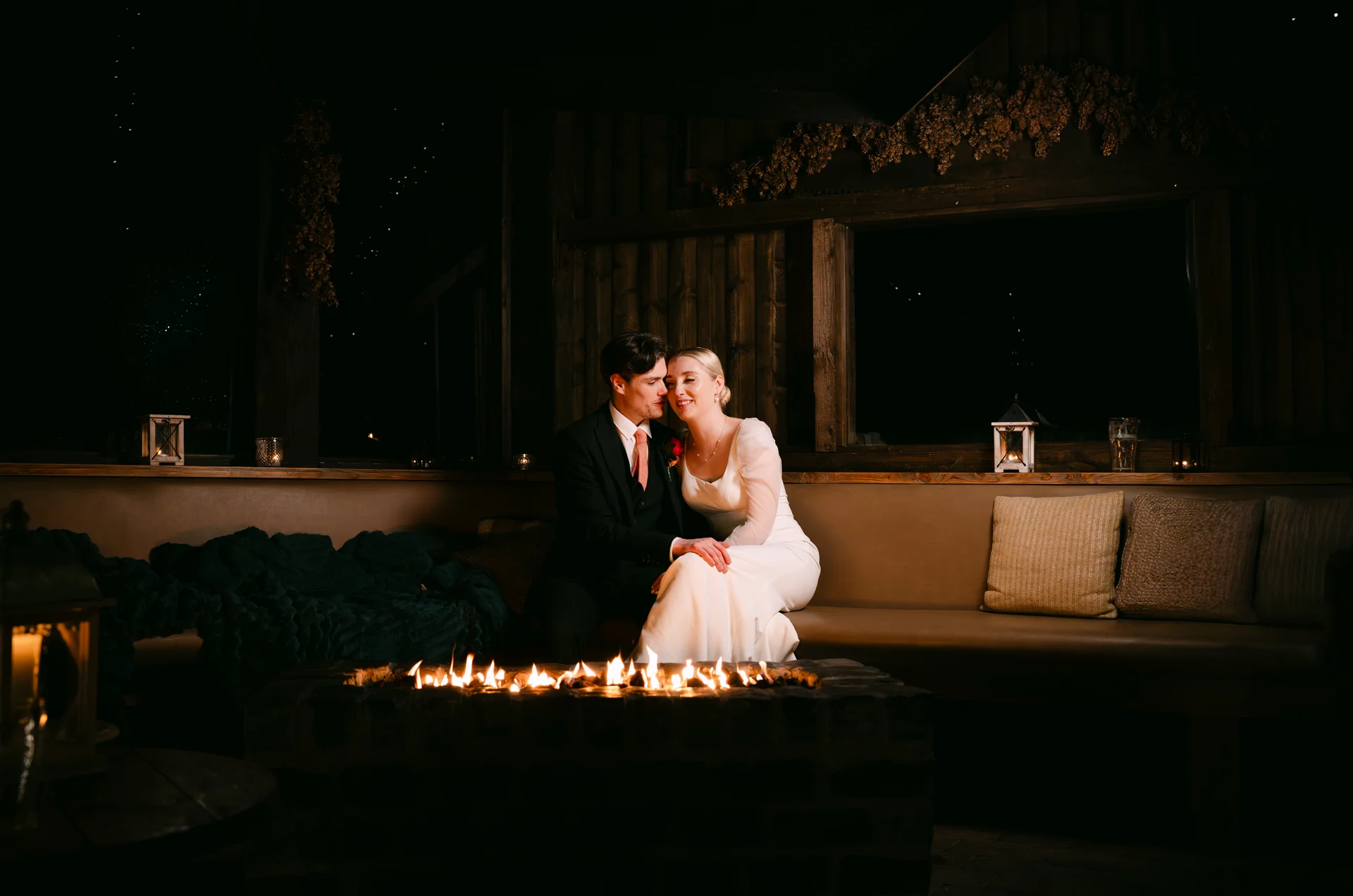A newlywed couple embraces lovingly next to a glowing brick firepit inside a rustic wood-panelled venue at night.