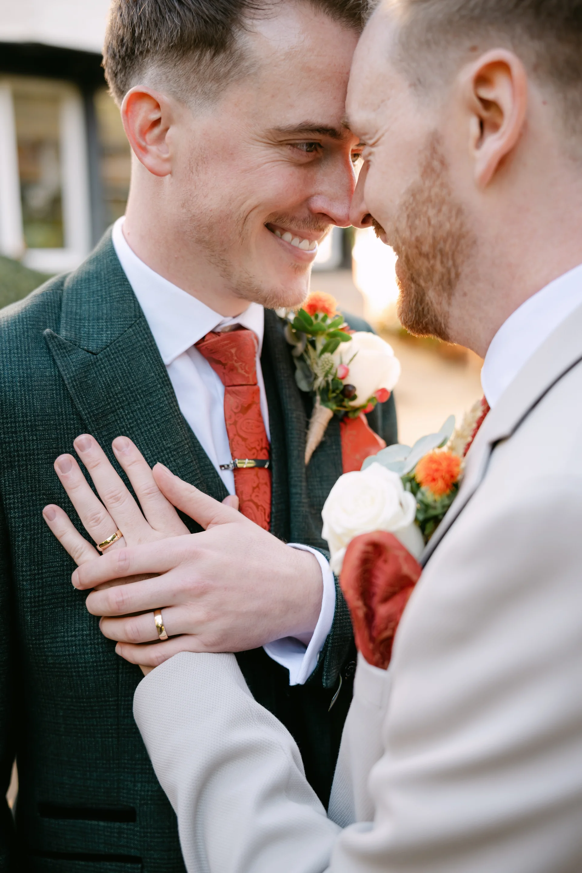 Two grooms share a tender nose-to-nose moment while smiling, showcasing their wedding bands and coordinated suits with floral buttonholes.