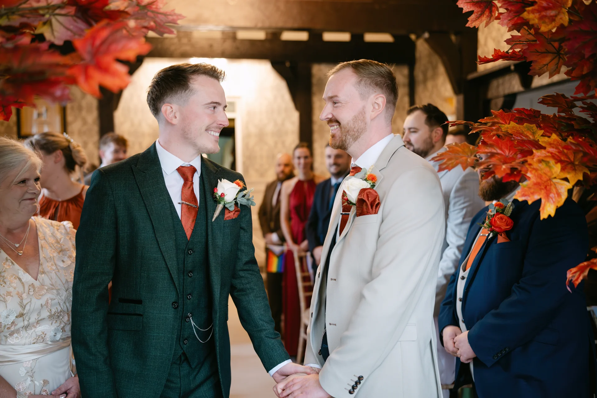 Two grooms share a loving look and hold hands during their autumn ceremony, framed by vibrant red and orange fall foliage.