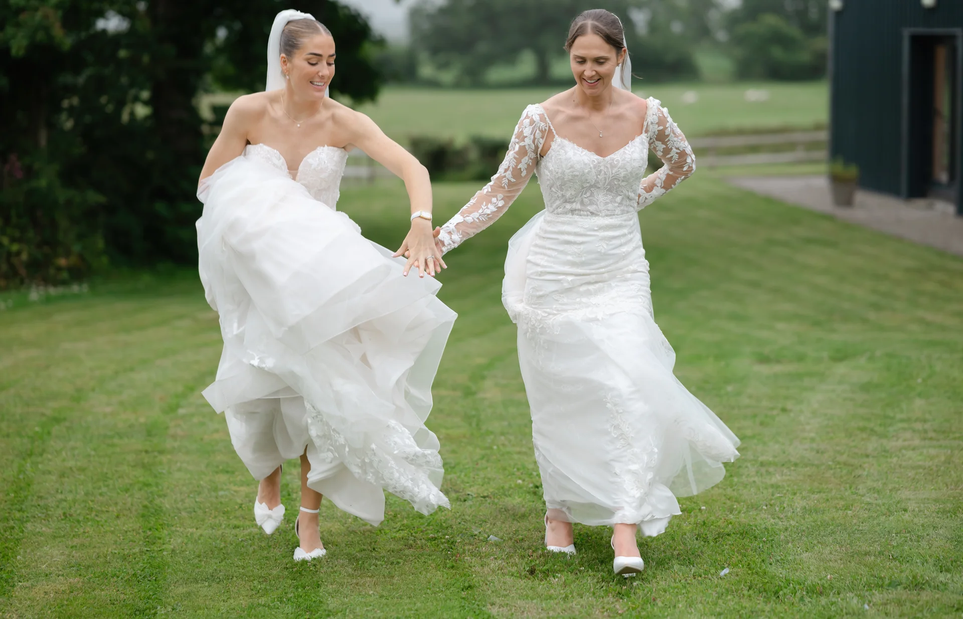 Two smiling brides in distinct wedding dresses hold hands as they run across a green lawn outdoors.