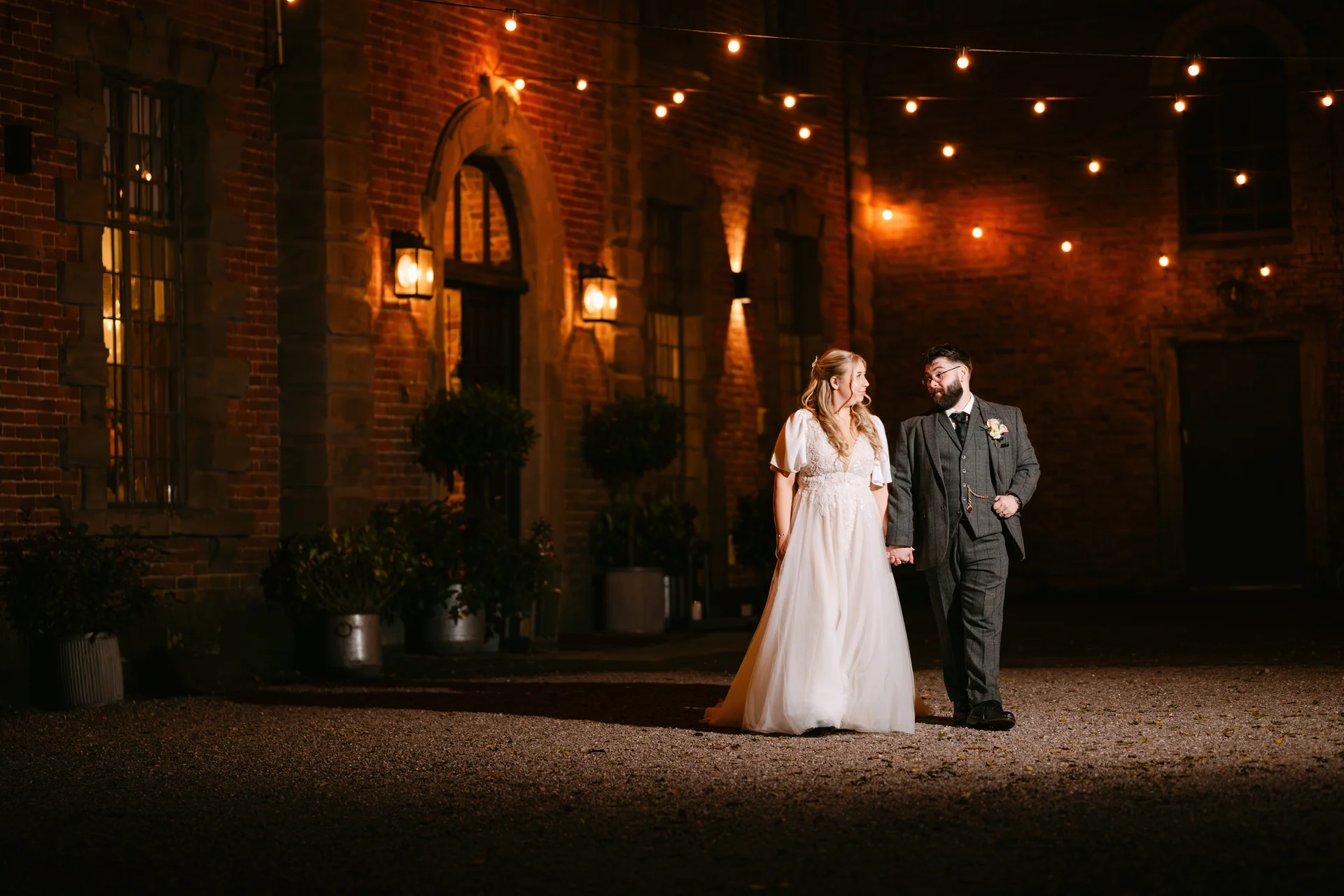 A newlywed couple walks hand in hand across a softly lit courtyard at night in front of a rustic brick building adorned with festoon lighting.