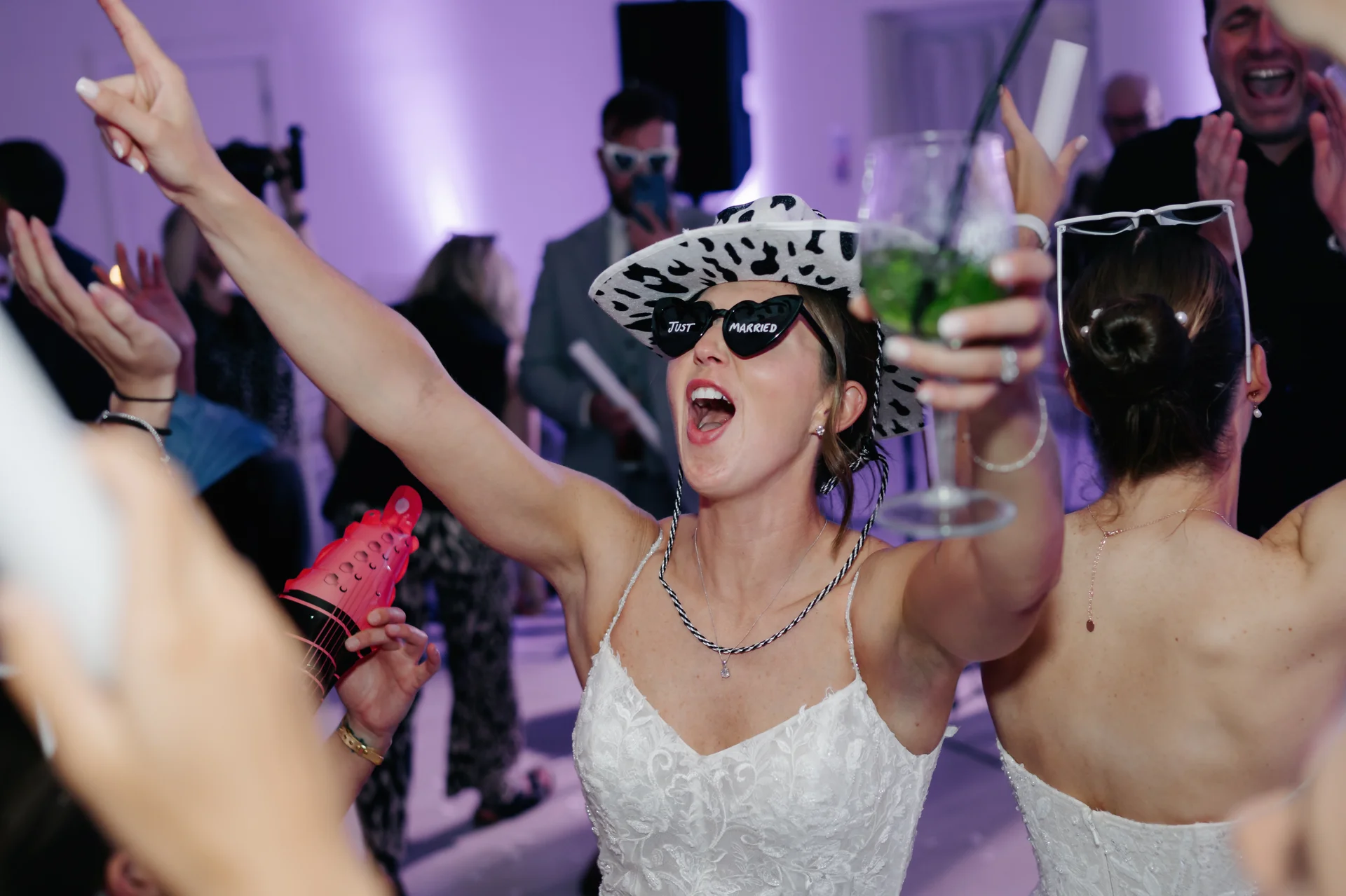 A joyful bride wearing a cowgirl hat and novelty sunglasses cheers on the packed dance floor while holding a cocktail glass during the evening reception.