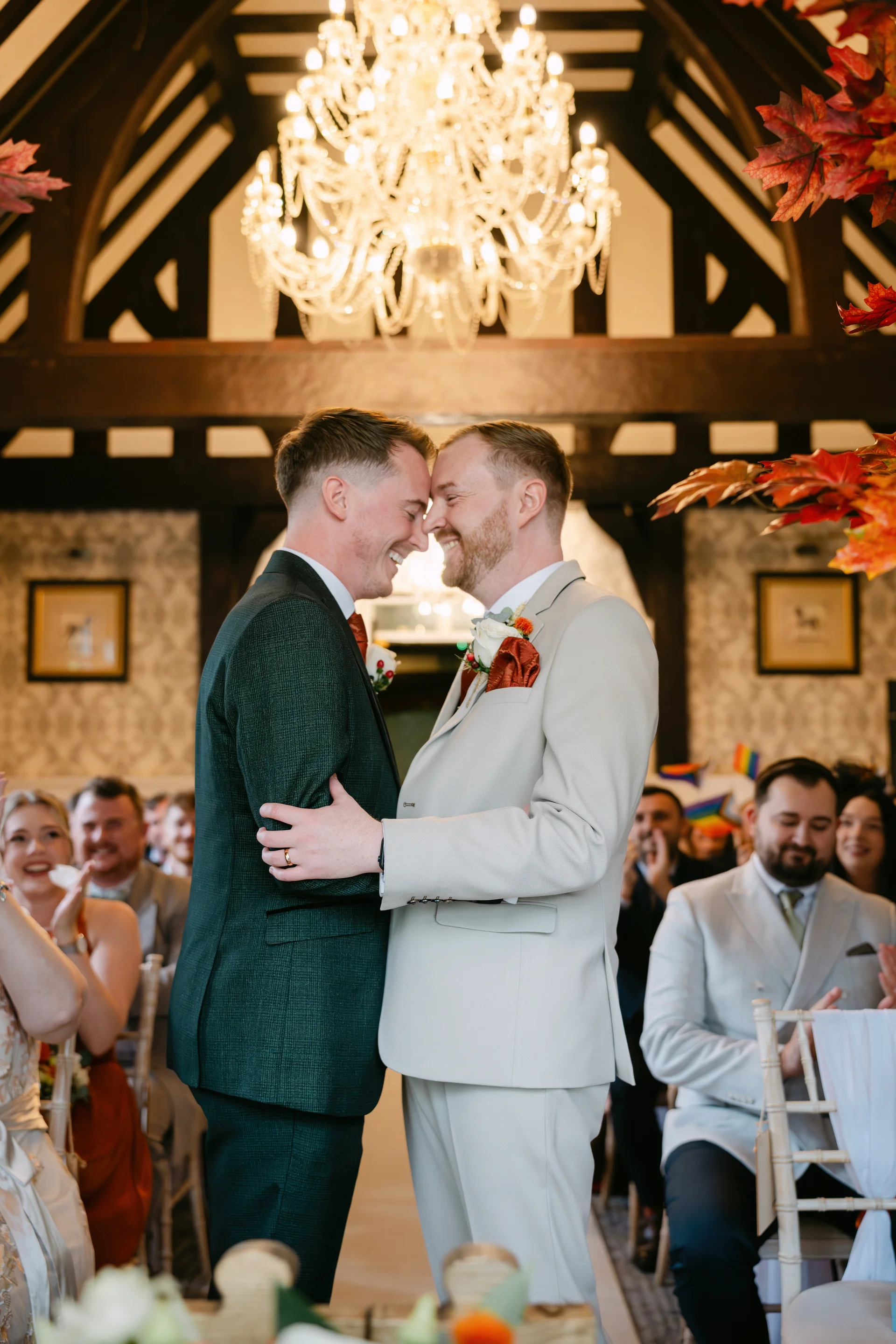 Two grooms share a tender forehead touch and smile happily at each other during their autumn barn wedding ceremony, surrounded by guests and overhead rustic decor.