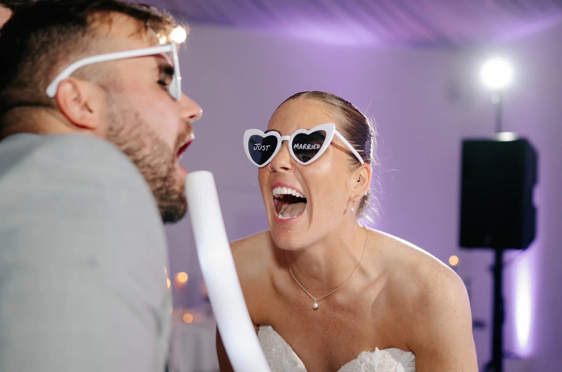 A joyful bride wearing heart-shaped sunglasses that read just married laughs excitedly on the dance floor alongside her partner during the evening reception.