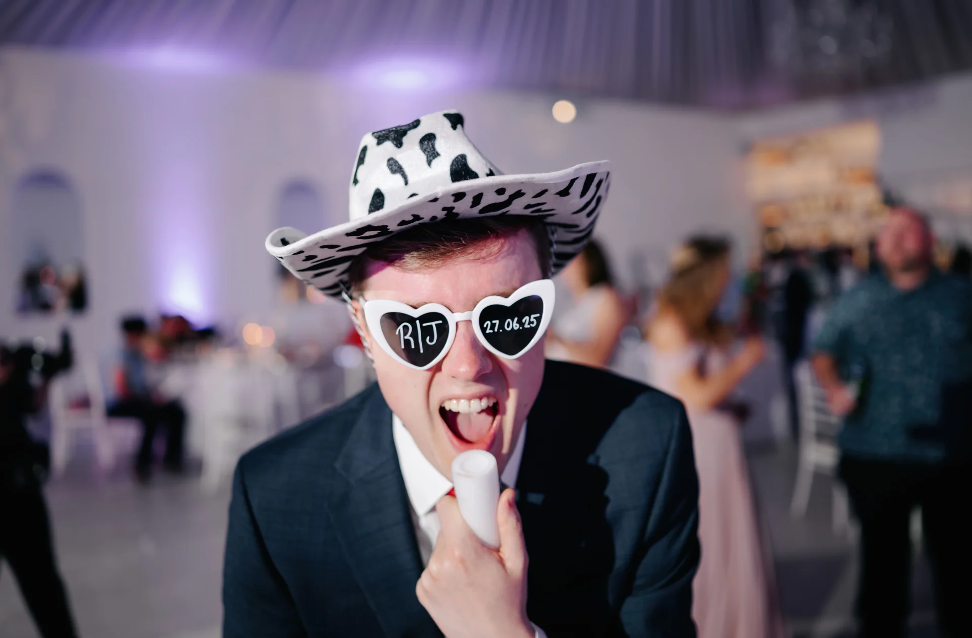 A wedding guest wearing a cow-print cowboy hat and heart-shaped sunglasses laughs enthusiastically on the dance floor during the evening reception.