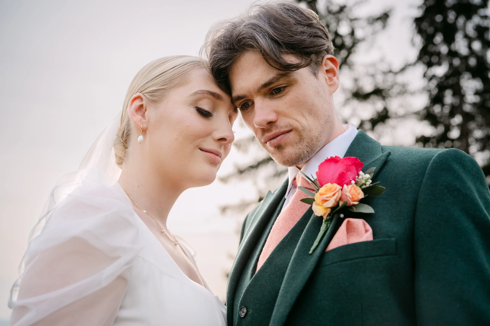 A close-up portrait of a newlywed couple standing outdoors, with the bride resting her forehead gently against the groom who wears an emerald green suit and floral buttonhole.