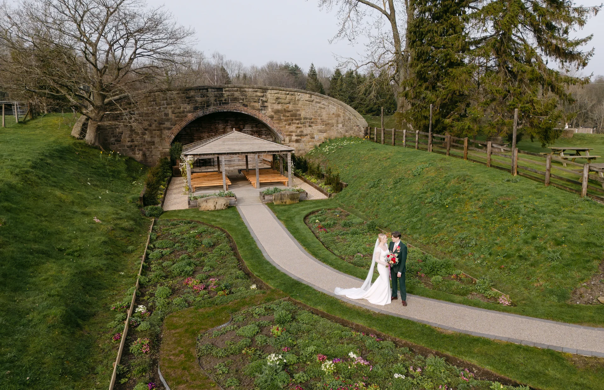 An aerial view captures a bride and groom standing on a winding path in a lush garden setting, with a stone bridge and outdoor ceremony pavilion in the background.