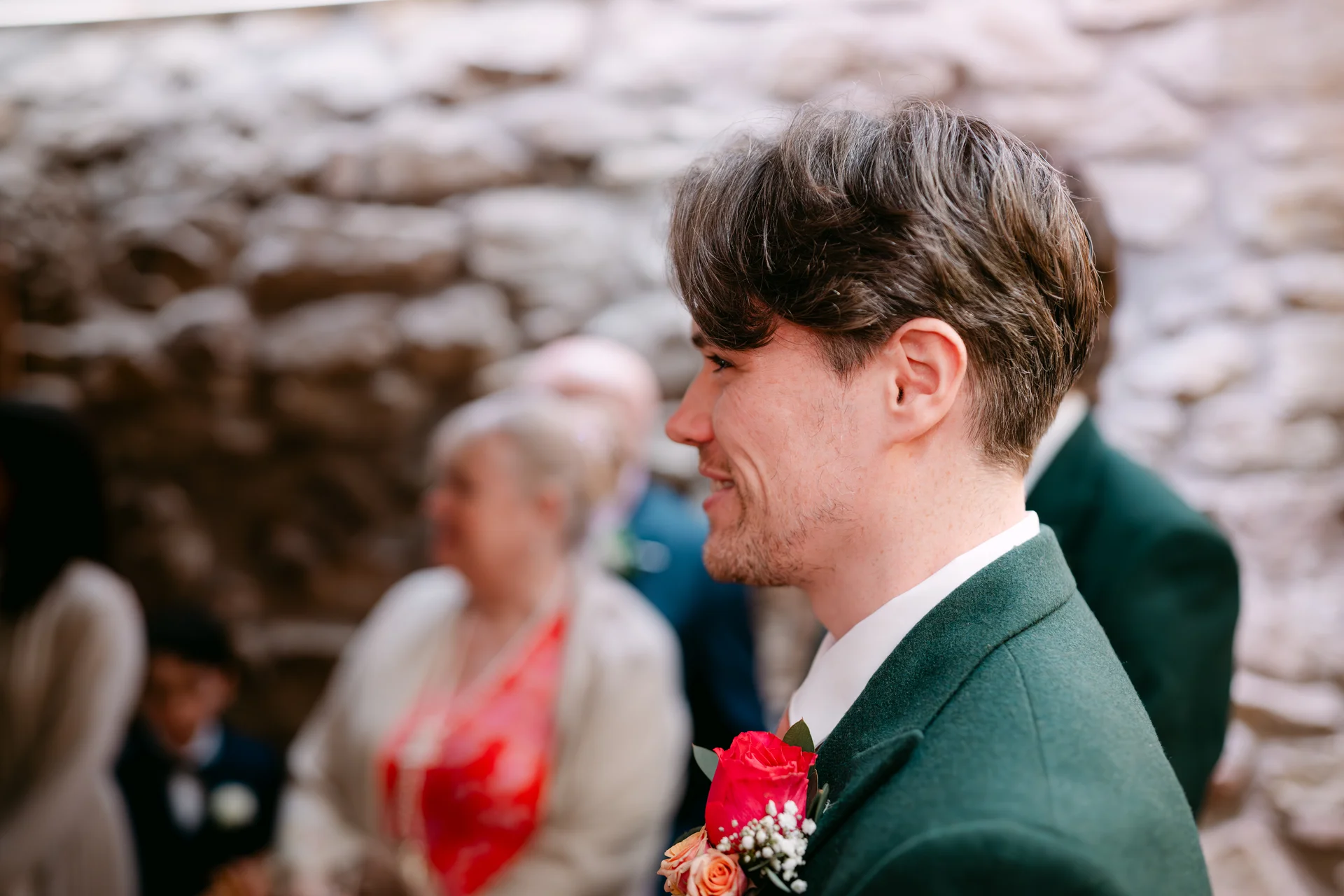 A groom wearing a green suit and boutonniere stands smiling in profile during a wedding ceremony inside a stone venue.