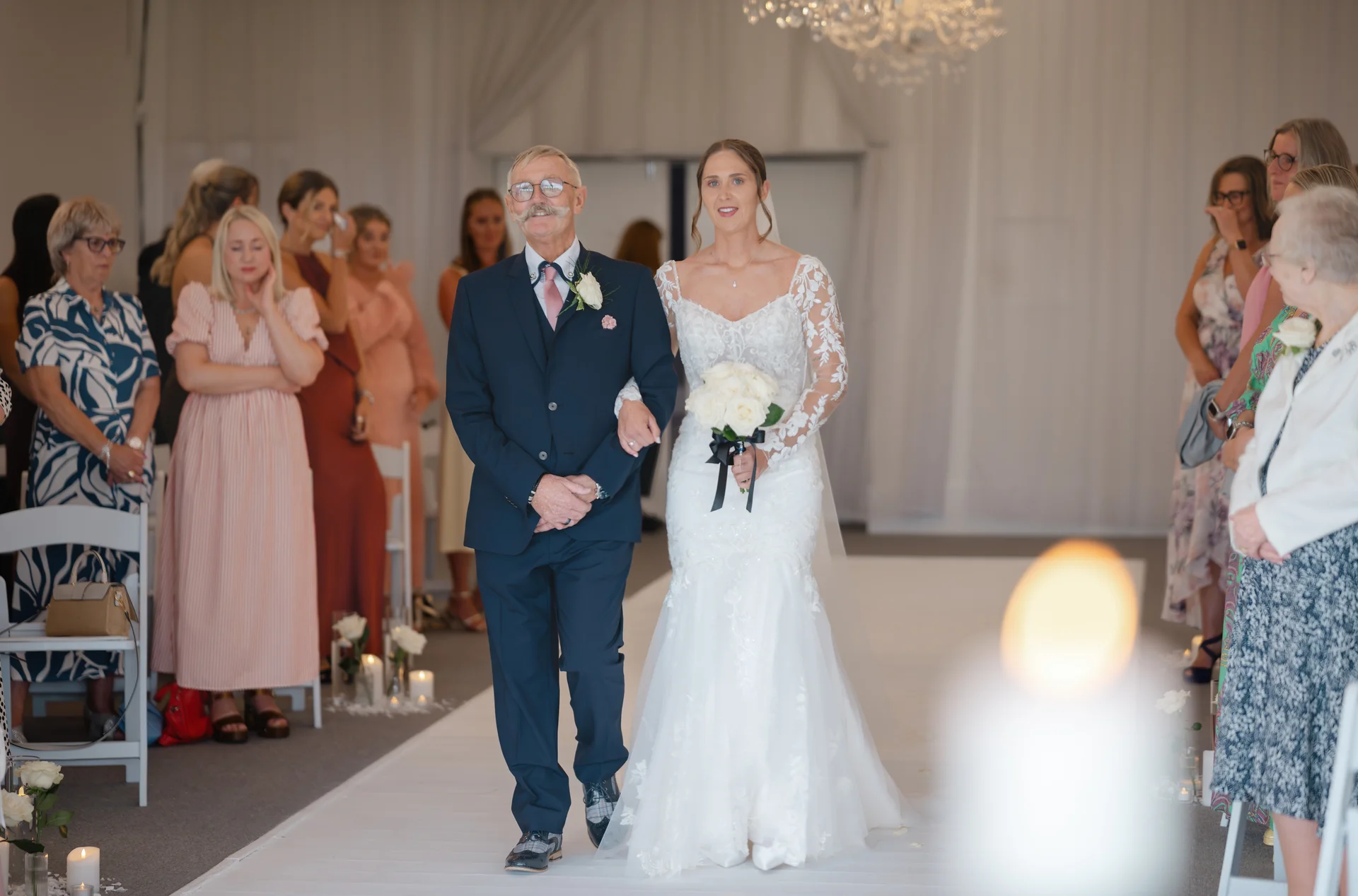 A proud father walks his smiling daughter in a long-sleeved lace wedding dress down the aisle while guests look on.