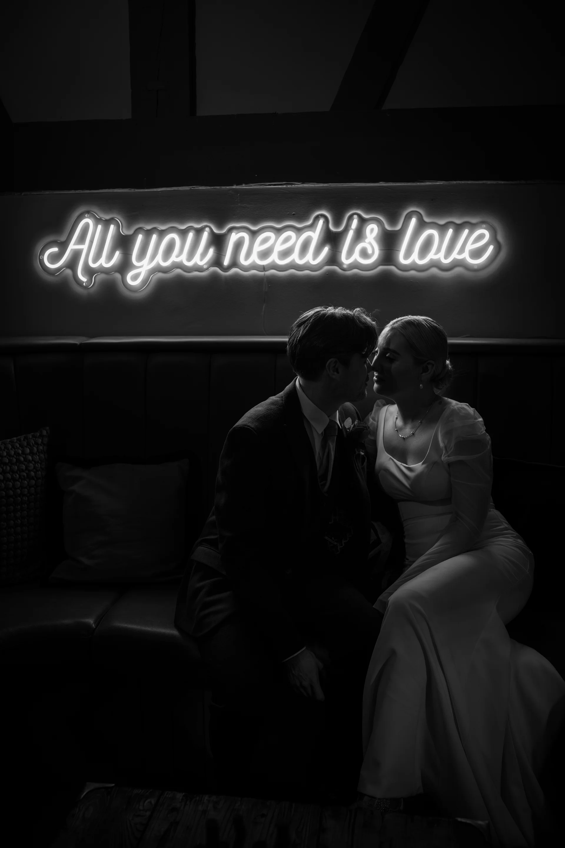 A newlywed couple sits closely together on a dark booth seating arrangement under a glowing neon sign reading All you need is love.