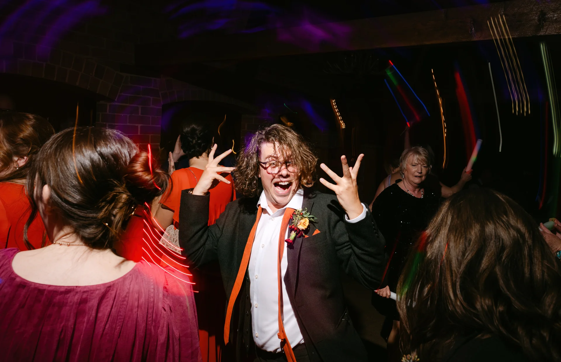 An energetic groom dances enthusiastically on a vibrant, colour-lit wedding dance floor while surrounded by partying guests.