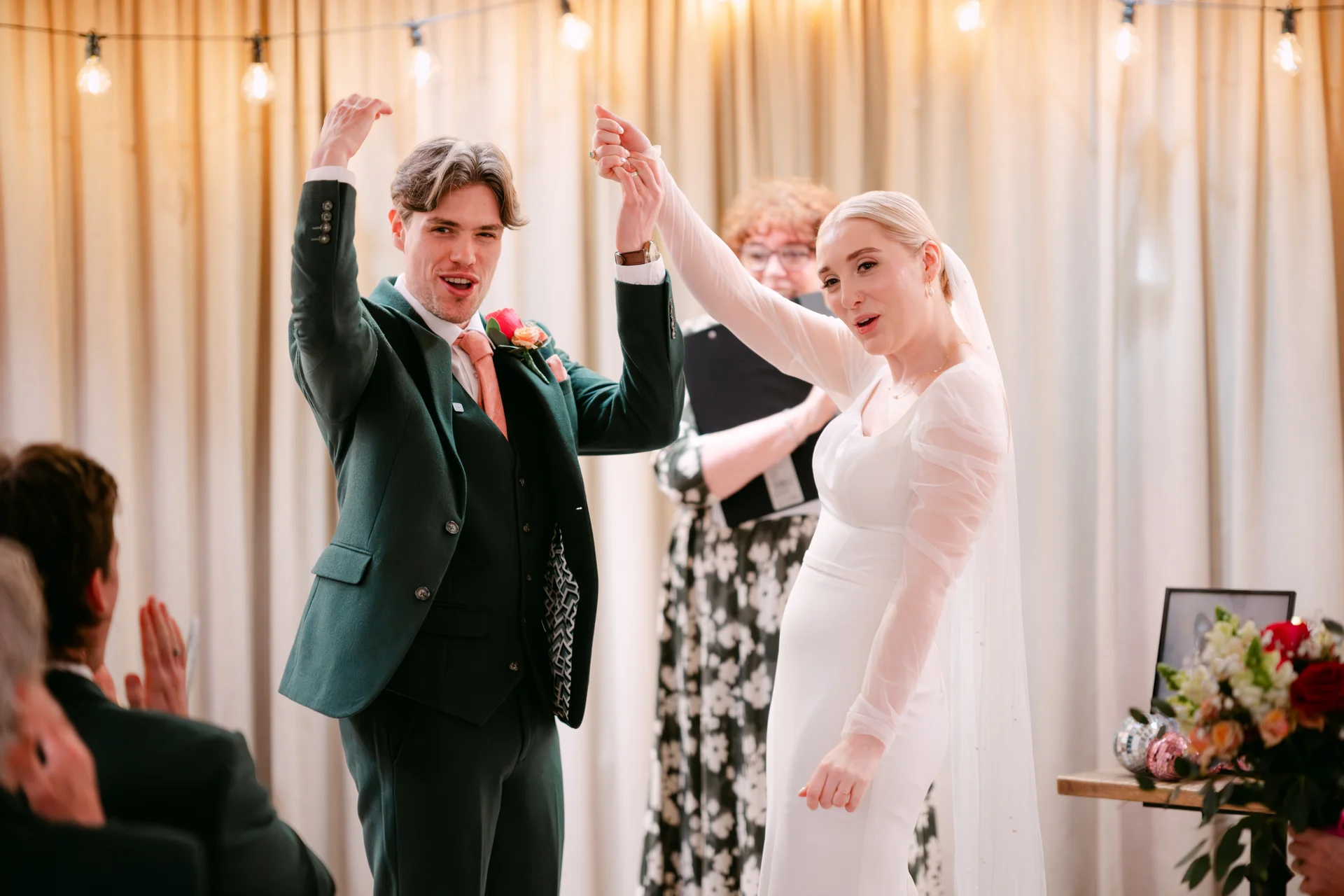 A newlywed couple celebrate at the altar during their civil ceremony, with the groom wearing a dark green suit and the bride in a white gown with sheer sleeves.