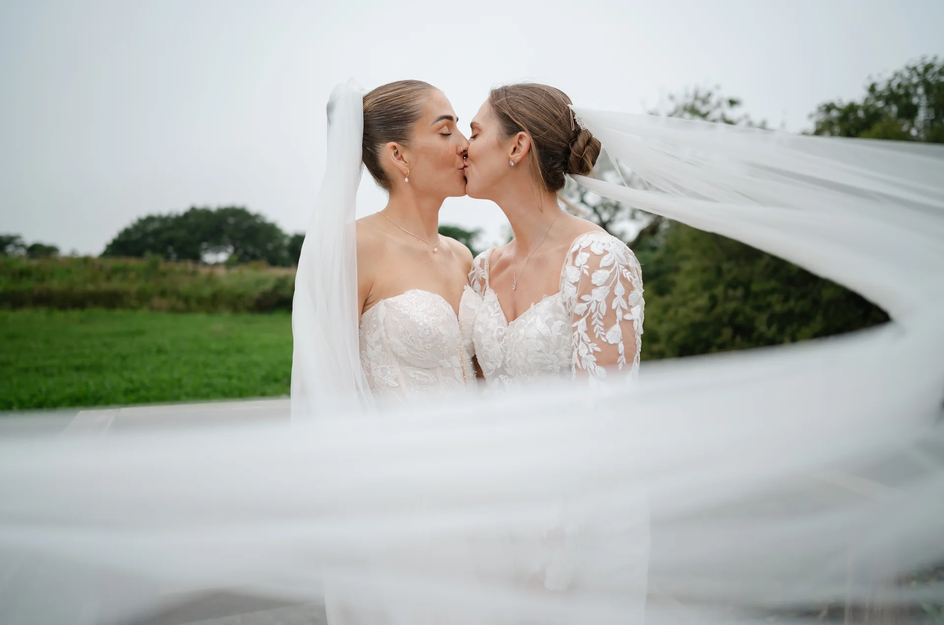 Two brides share a tender kiss outdoors with a dramatic veil blowing across the foreground against an overcast landscape.